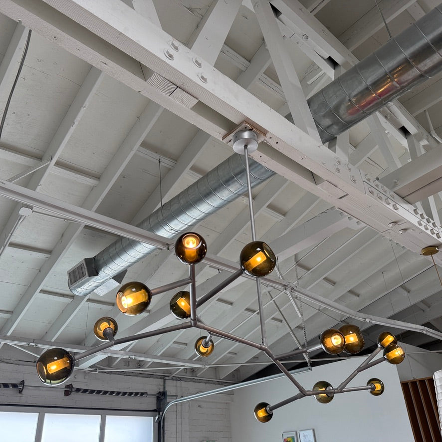 The Pouenat Last Night Branche Chandelier by Pouenat, featuring mouth-blown glass spheres, hangs beneath a white exposed beam ceiling with visible ductwork—an ideal statement for modern industrial interiors.