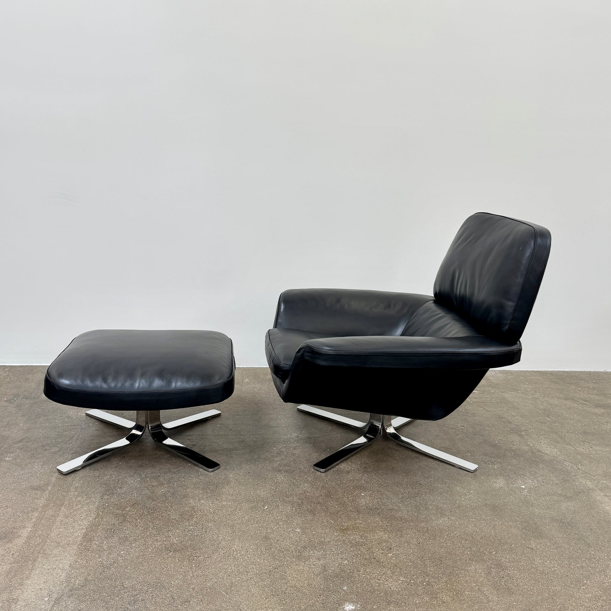 A Minotti Blake Lounge Chair & Ottoman by Minotti in black leather with chrome swivel bases, positioned on a bare concrete floor against a plain light-colored wall.