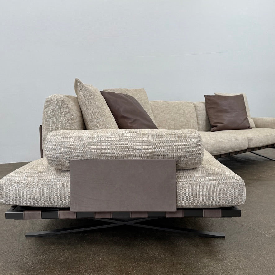 The Driade Fenix sofa by Driade is a beige L-shaped modular design with three seat cushions, black legs, and four throw pillows—two brown, two beige—highlighting its architectural lightness against a plain white backdrop.
