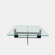The Minotti Ellis Coffee Table Clear Glass by Minotti features a square clear glass top and a sculptural black metal base, elegantly positioned on a brown concrete floor against a plain white wall.