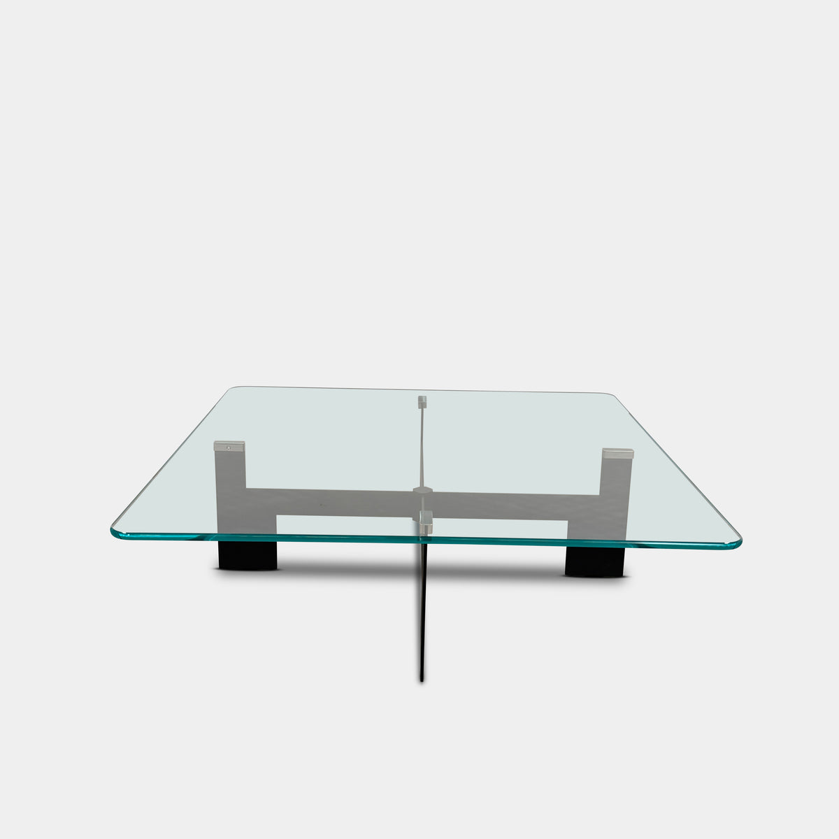 The Minotti Ellis Coffee Table Clear Glass by Minotti features a square clear glass top and a sculptural black metal base, elegantly positioned on a brown concrete floor against a plain white wall.