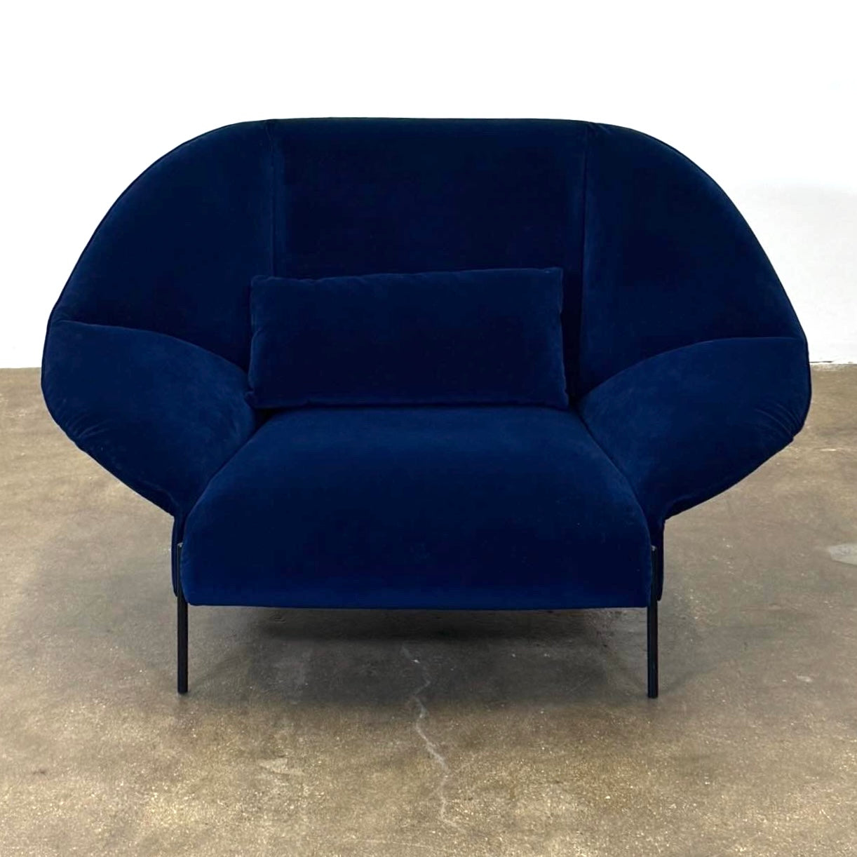 The Ligne Roset Paipai Armchair by Ligne Roset features a navy blue velvet finish, curved cocoon-like seat, matching rectangular cushion, slim black legs, and premium foam construction, displayed against a plain background.