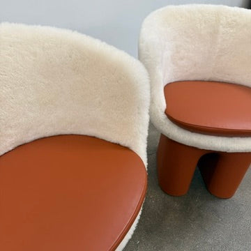 The Louis Vuitton Dolls Fur by Raw Edges is a modern chair with a curved white fluffy leather and fur seat, two short cylindrical brown legs, and a matching brown base, designed by Louis Vuitton and set against a plain background.