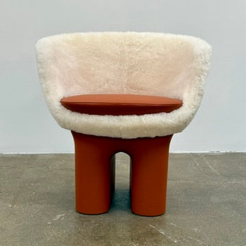 The Louis Vuitton Dolls Fur by Raw Edges is a modern chair with a curved white fluffy leather and fur seat, two short cylindrical brown legs, and a matching brown base, designed by Louis Vuitton and set against a plain background.