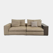 The Flexform Groundpiece Sofa With Leather Shelving by Flexform is a beige two-seater featuring four large cushions, one small beige cushion, and a dark leather shelf attached to the right armrest for sleek modern seating.