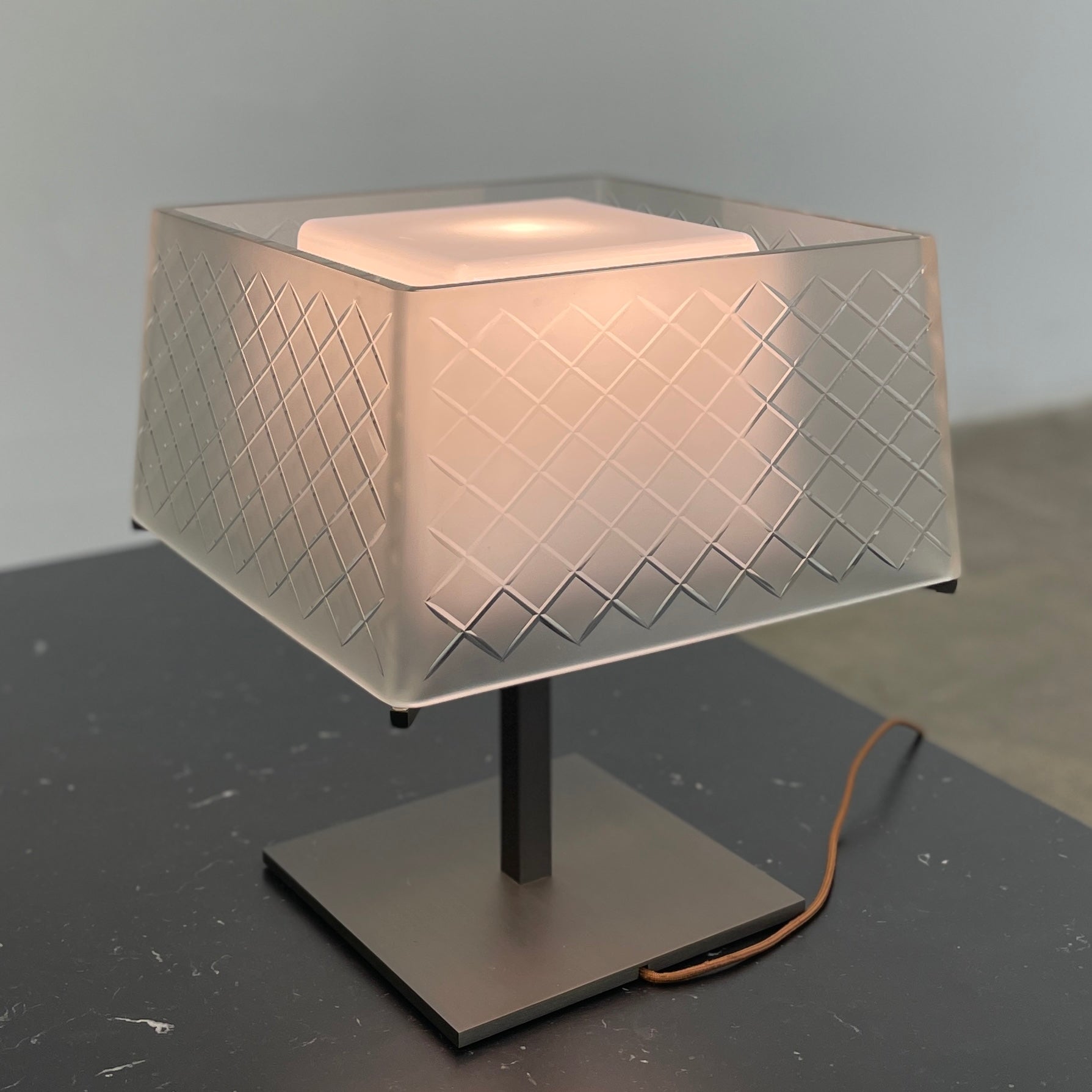 Introducing the Flos Metropolitan Table Light by Flos: a chic table lamp with a rectangular shade adorned with a diamond pattern, gracefully positioned on a square base.