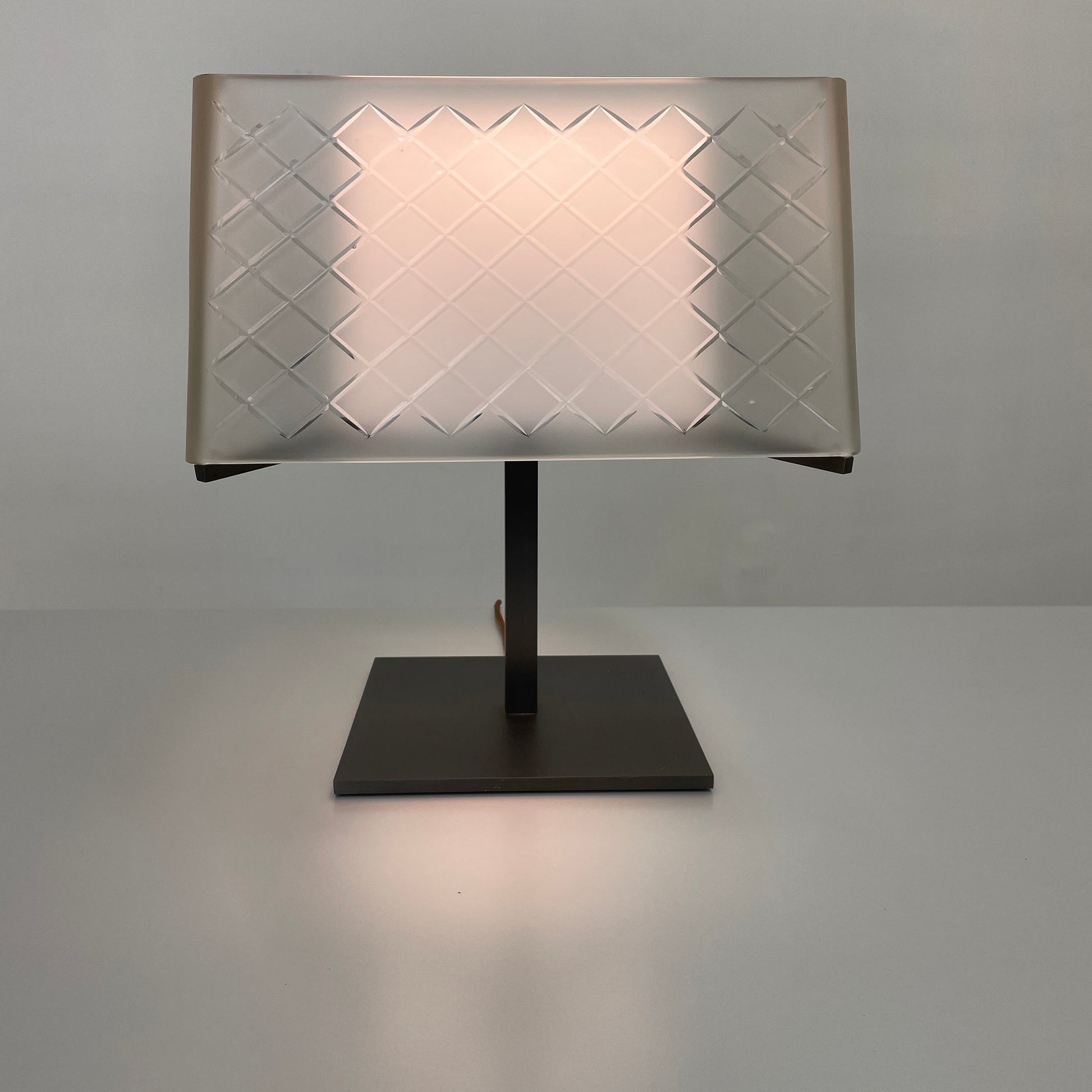 Introducing the Flos Metropolitan Table Light by Flos: a chic table lamp with a rectangular shade adorned with a diamond pattern, gracefully positioned on a square base.