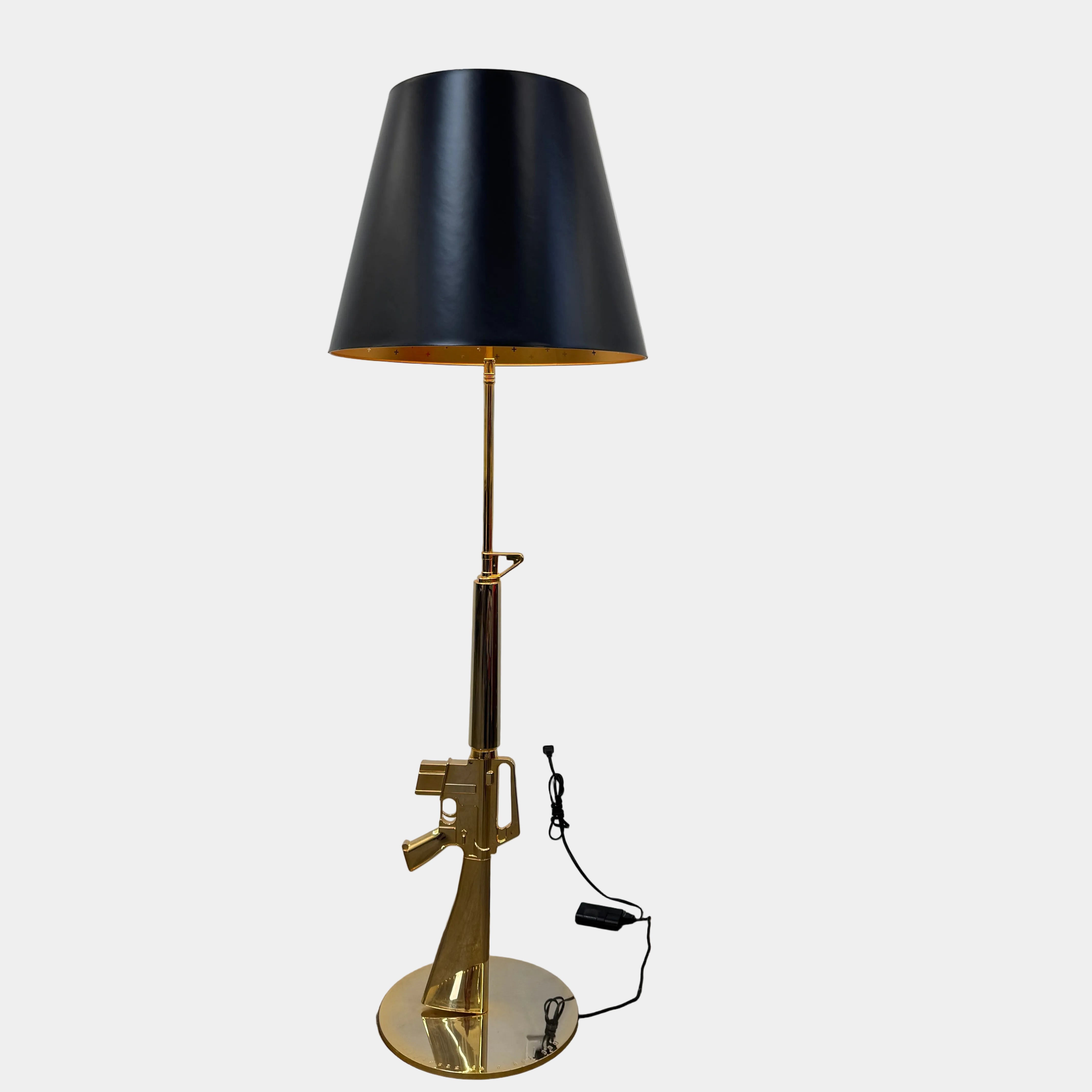 The Flos Guns Lounge Floor Lamp Gold by Flos features a shiny gold rifle-shaped base and a black lampshade, inspired by Philippe Starck, standing against a plain white background.