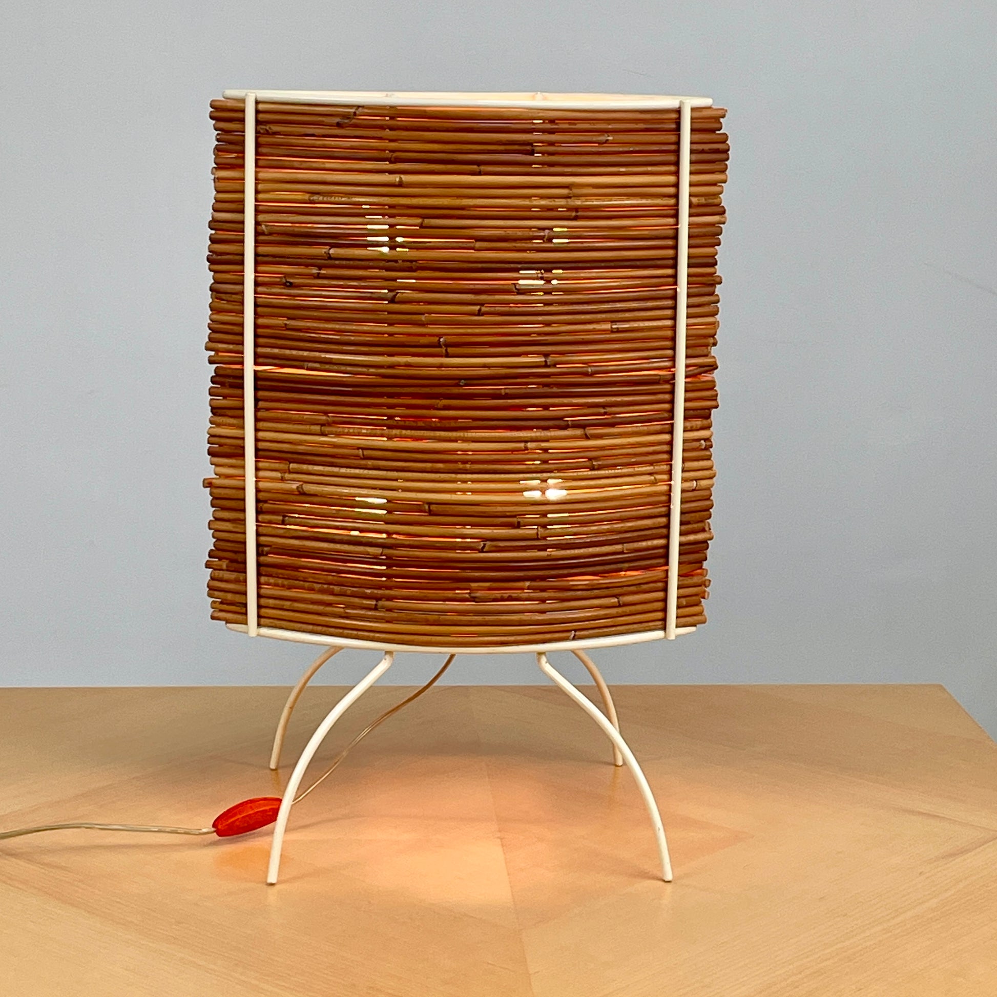 The Vintage Fontana Arte Bambú Table Light by Fontana Arte showcases a woven rattan circular shade atop a sleek white metal tripod base, making it an elegant addition to a wooden table. This lamp enhances your home's interior lighting with its natural charm and unique design.