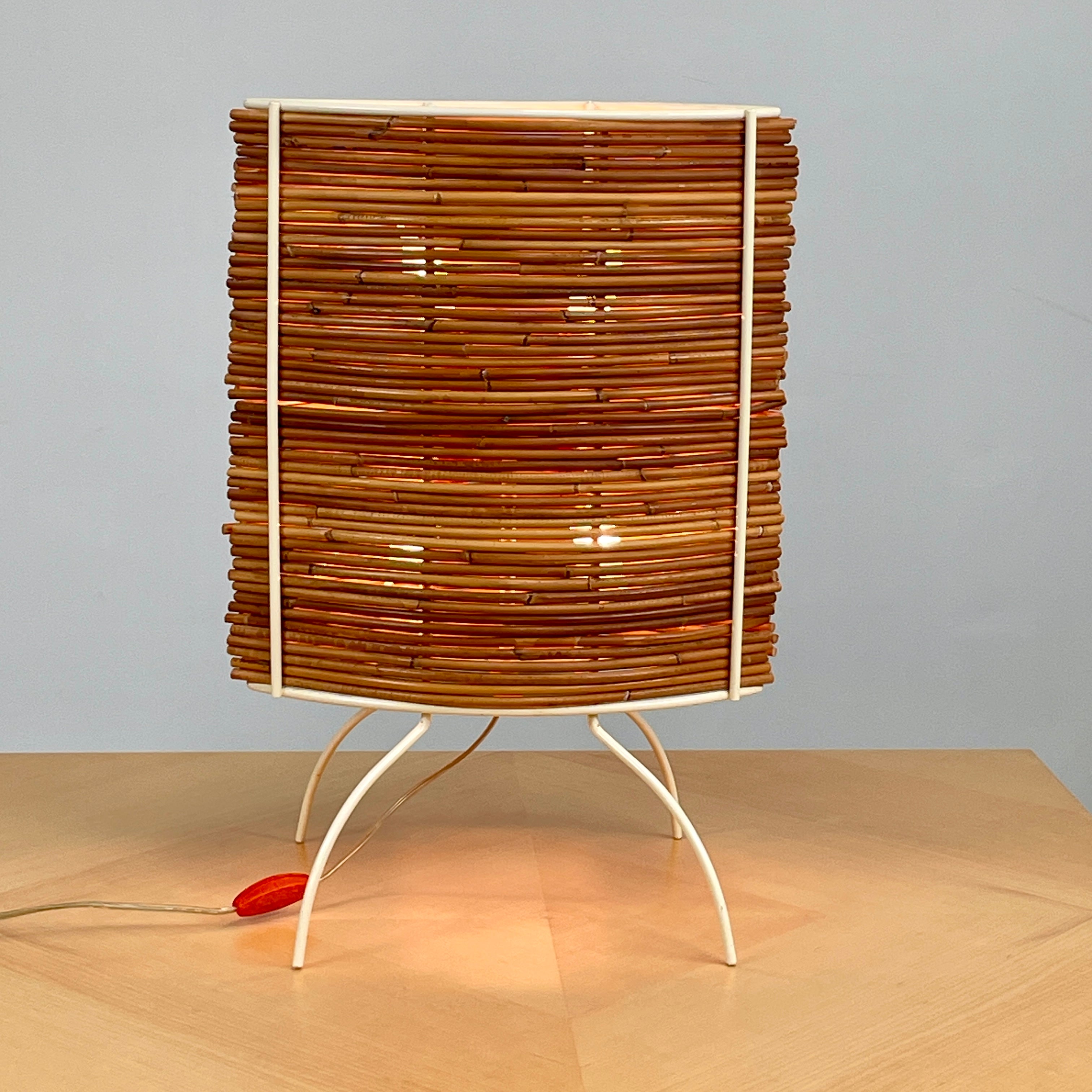 The Vintage Fontana Arte Bambú Table Light by Fontana Arte showcases a woven rattan circular shade atop a sleek white metal tripod base, making it an elegant addition to a wooden table. This lamp enhances your home's interior lighting with its natural charm and unique design.