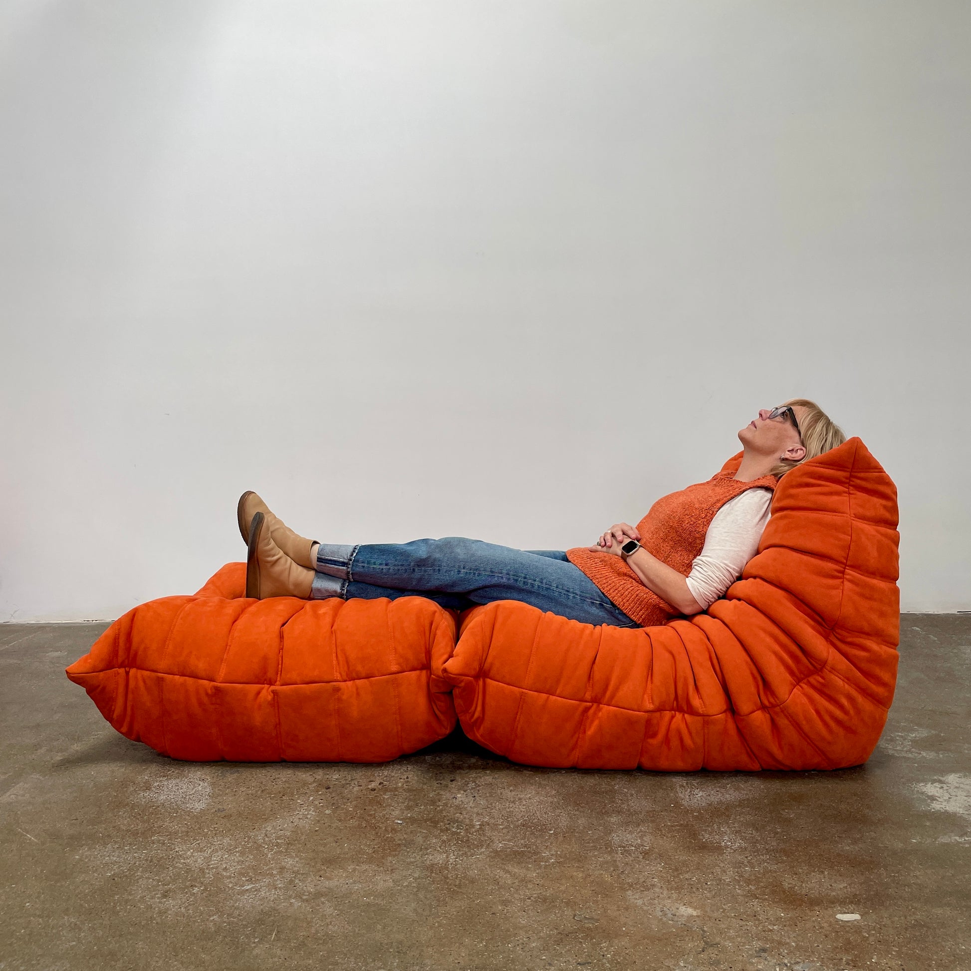 A Ligne Roset Togo Fireside Chair and Ottoman in orange, echoing the iconic design by Michel Ducaroy, is displayed against a pristine white backdrop.