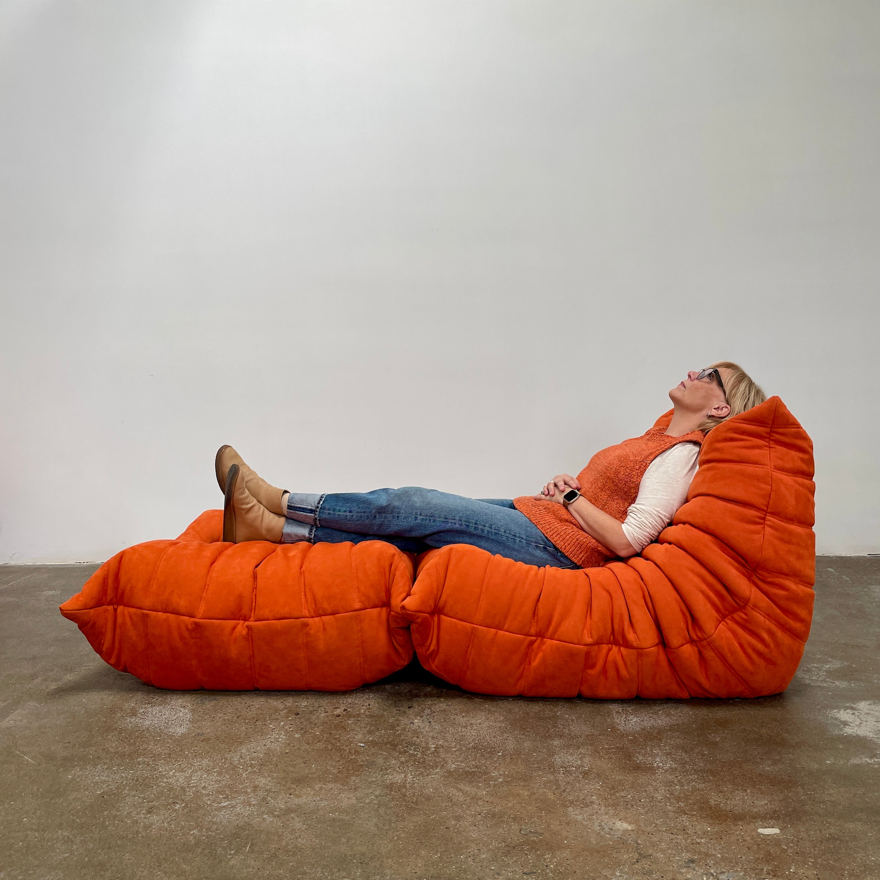 A Ligne Roset Togo Fireside Chair and Ottoman in orange, echoing the iconic design by Michel Ducaroy, is displayed against a pristine white backdrop.