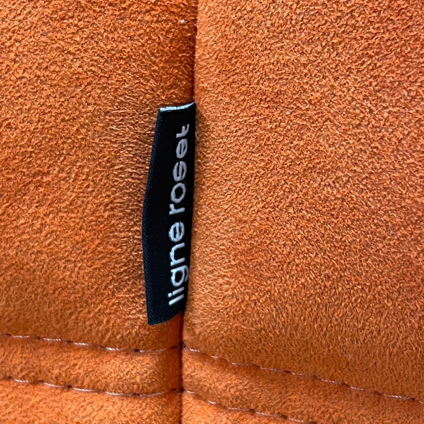 A Ligne Roset Togo Fireside Chair and Ottoman in orange, echoing the iconic design by Michel Ducaroy, is displayed against a pristine white backdrop.