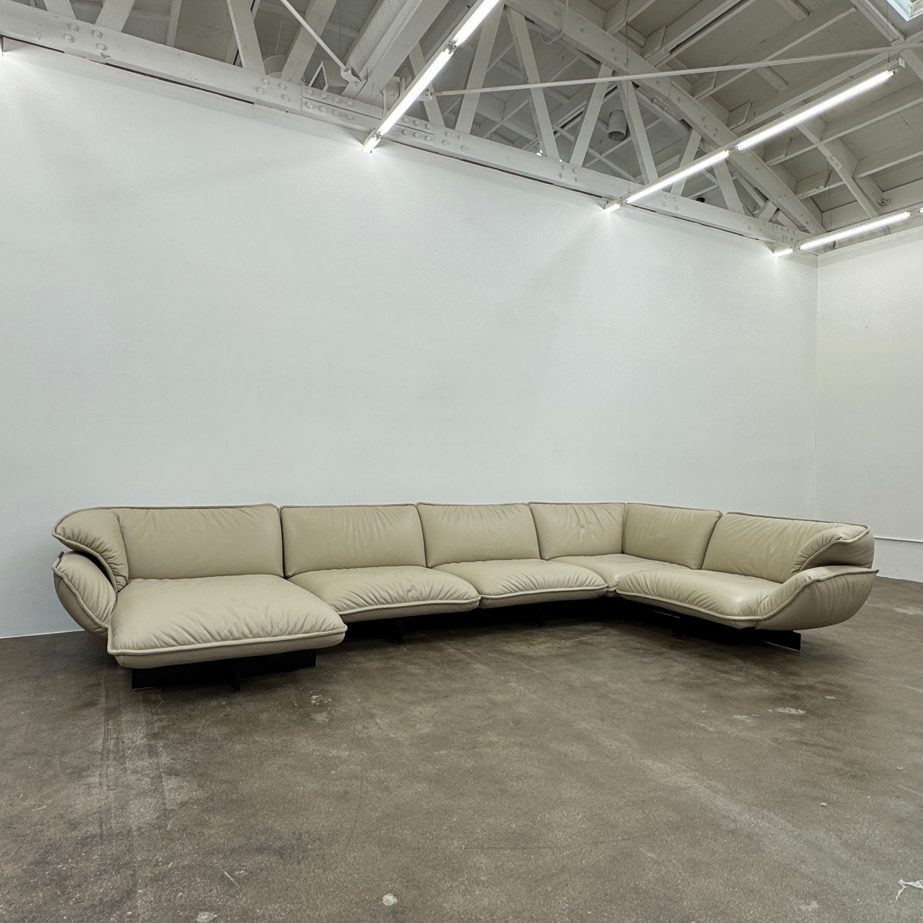 The Cassina Superbeam 5 Piece Leather Sectional in beige, featuring plush cushions, is arranged in a spacious room with white walls, exposed Superbeam ceiling, and overhead lighting.