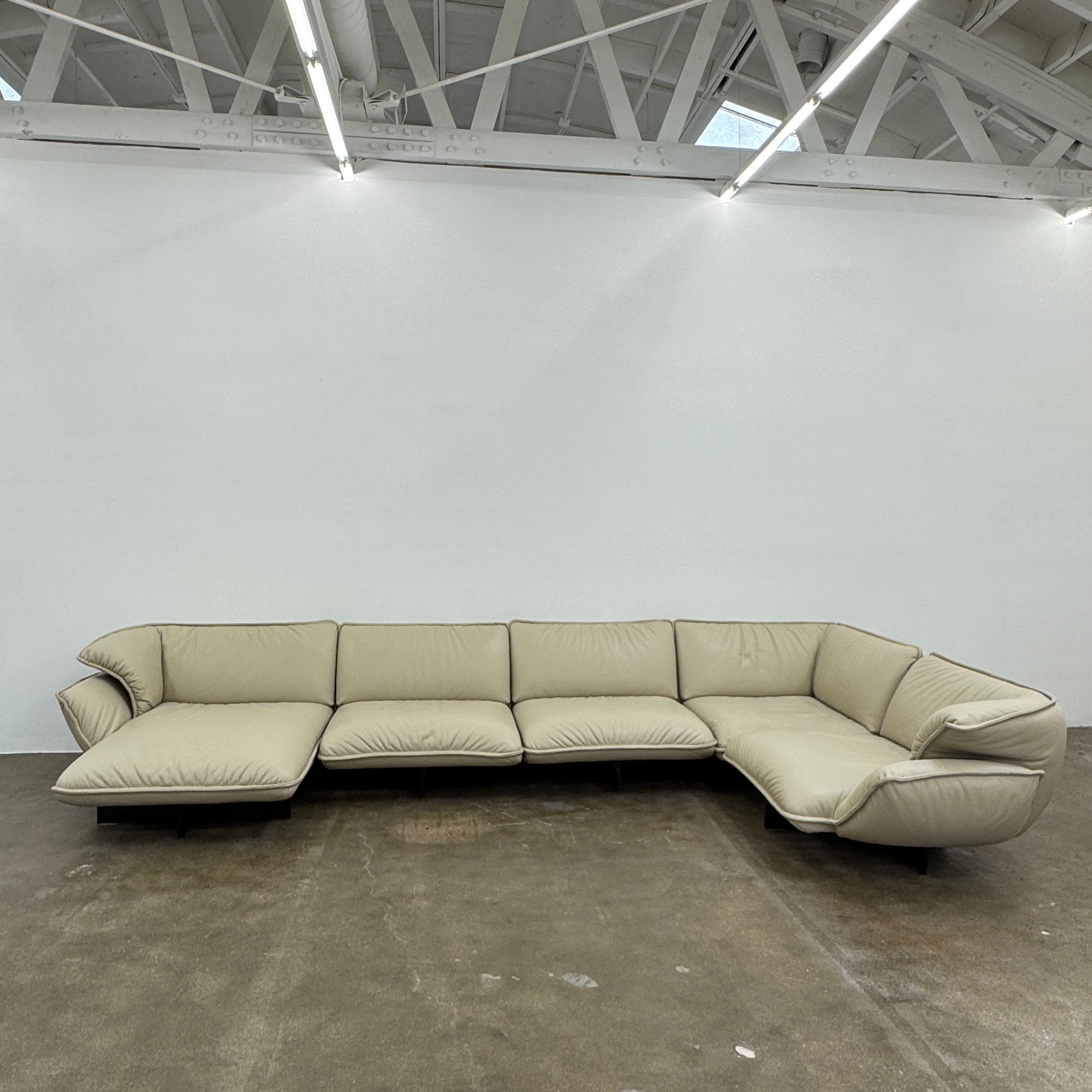 The Cassina Superbeam 5 Piece Leather Sectional in beige, featuring plush cushions, is arranged in a spacious room with white walls, exposed Superbeam ceiling, and overhead lighting.