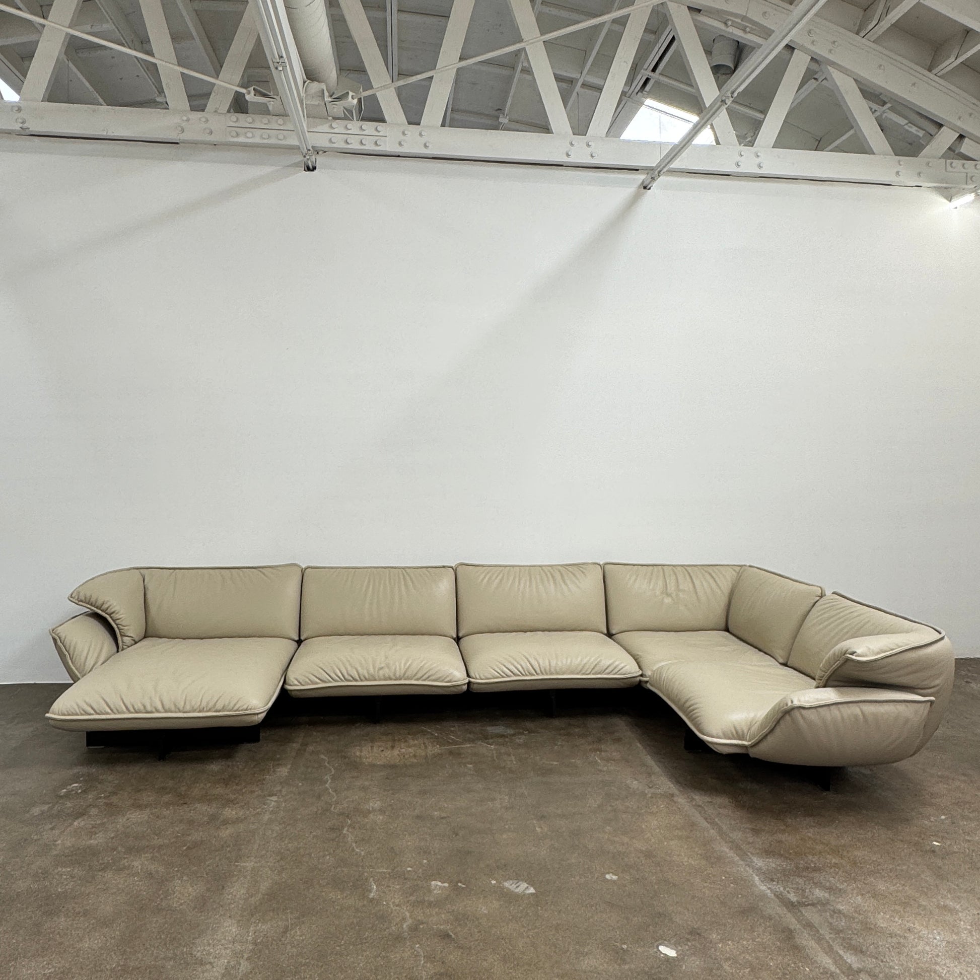 The Cassina Superbeam 5 Piece Leather Sectional in beige, featuring plush cushions, is arranged in a spacious room with white walls, exposed Superbeam ceiling, and overhead lighting.
