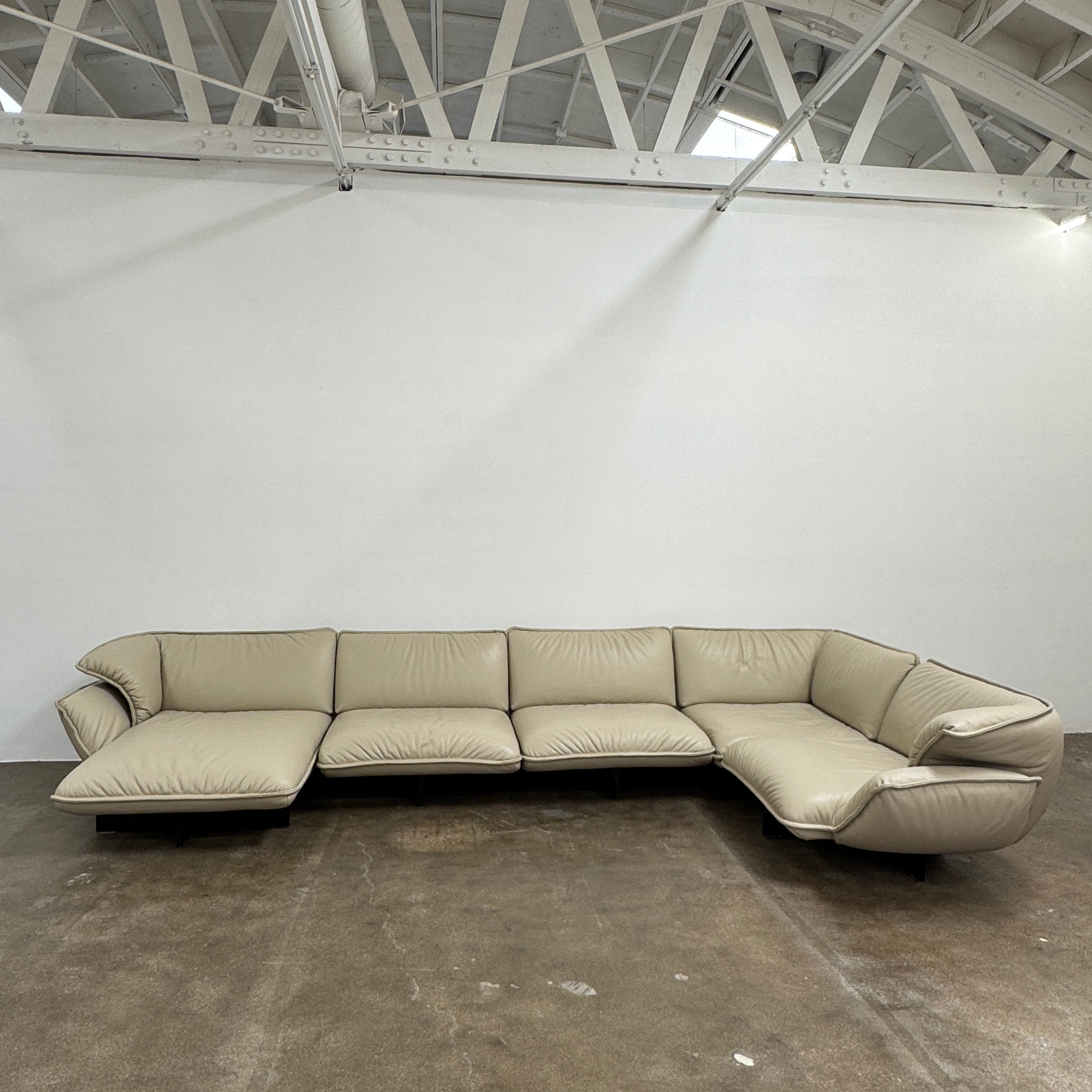 The Cassina Superbeam 5 Piece Leather Sectional in beige, featuring plush cushions, is arranged in a spacious room with white walls, exposed Superbeam ceiling, and overhead lighting.