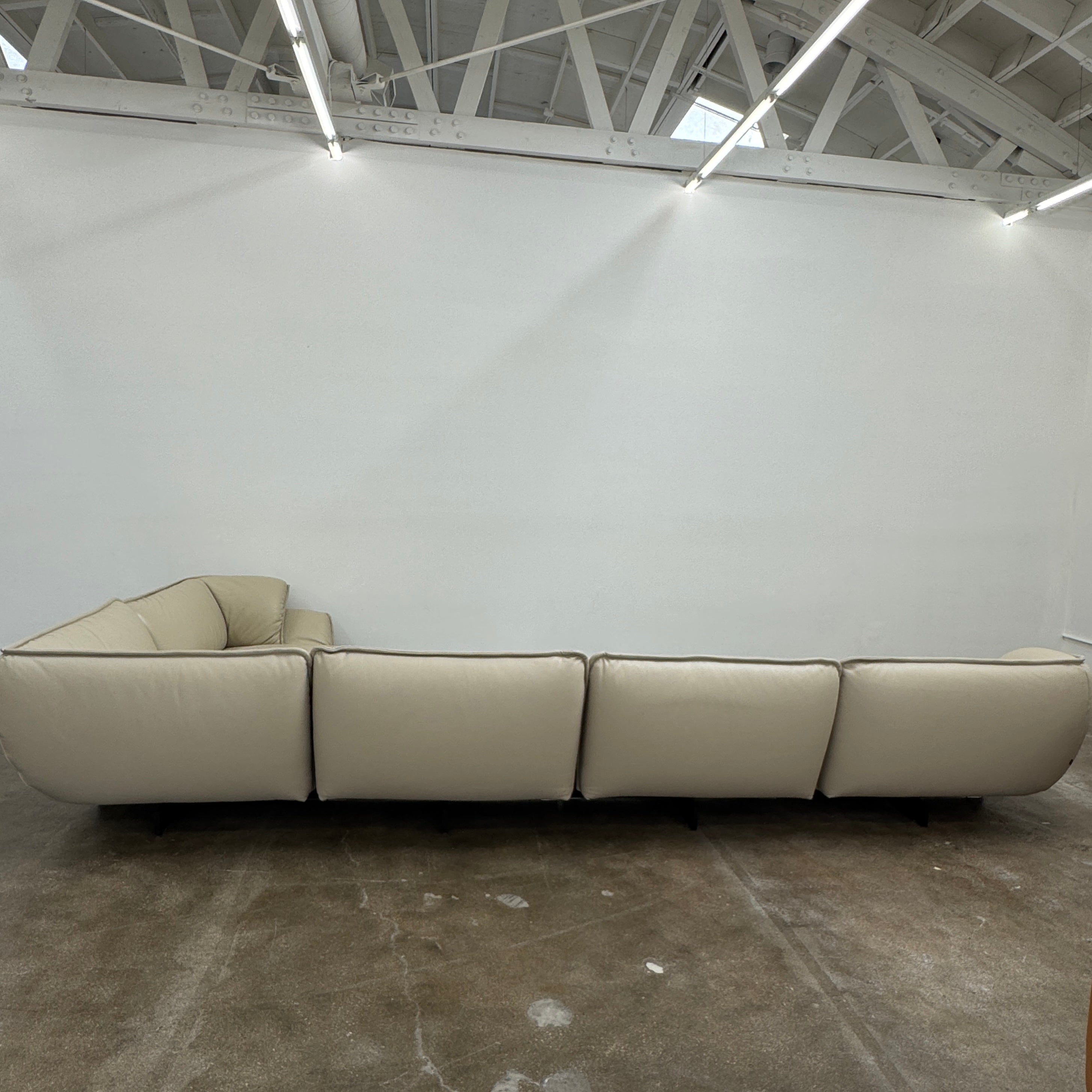 The Cassina Superbeam 5 Piece Leather Sectional in beige, featuring plush cushions, is arranged in a spacious room with white walls, exposed Superbeam ceiling, and overhead lighting.