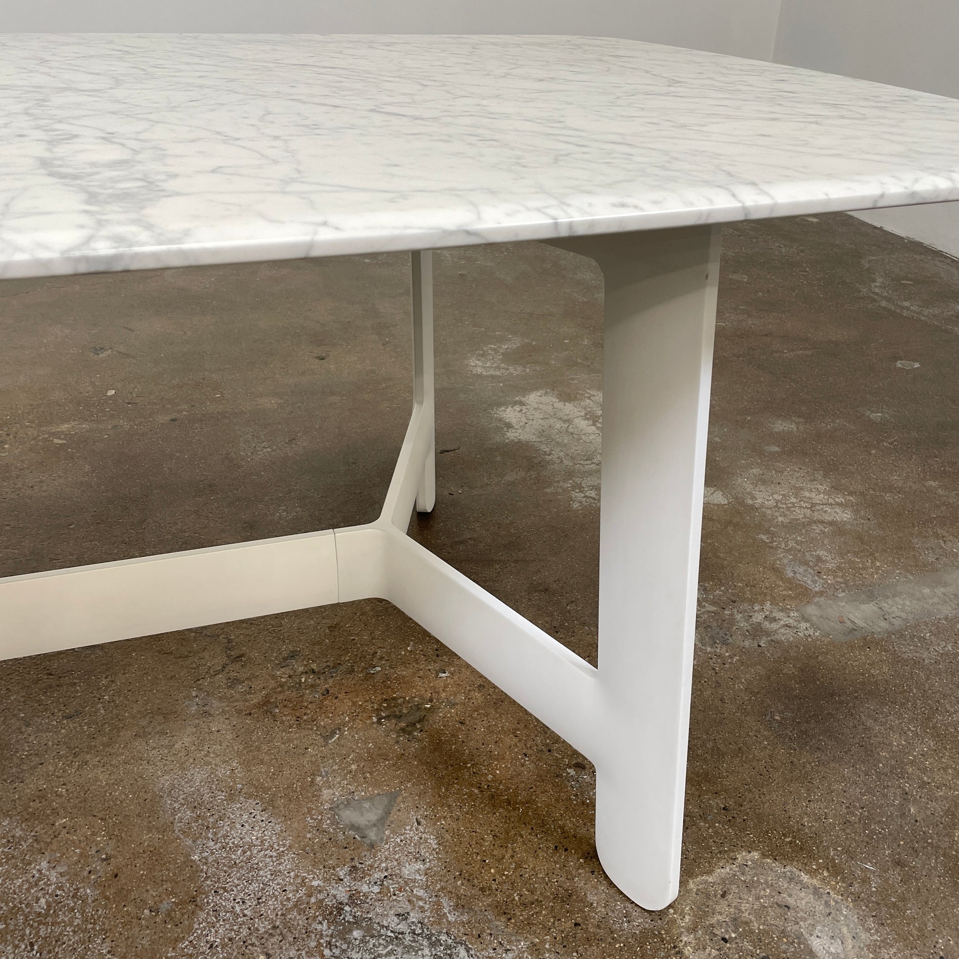 On a concrete floor against a plain white wall stands the B&B Italia Matte Carrara Marble Alex Dining Table, showcasing contemporary design with its long, rectangular form and white base, adding both style and sophistication to the space.