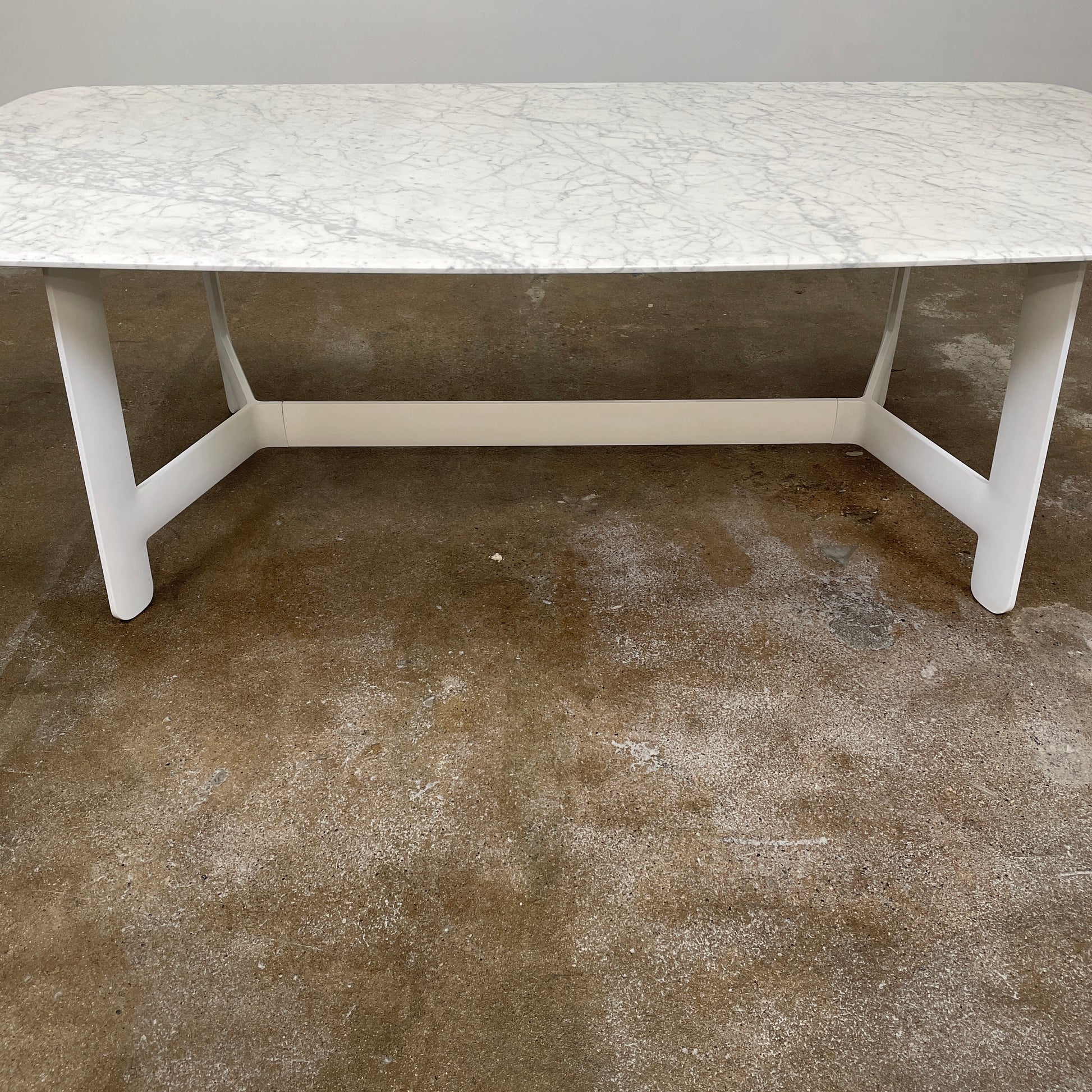 On a concrete floor against a plain white wall stands the B&B Italia Matte Carrara Marble Alex Dining Table, showcasing contemporary design with its long, rectangular form and white base, adding both style and sophistication to the space.