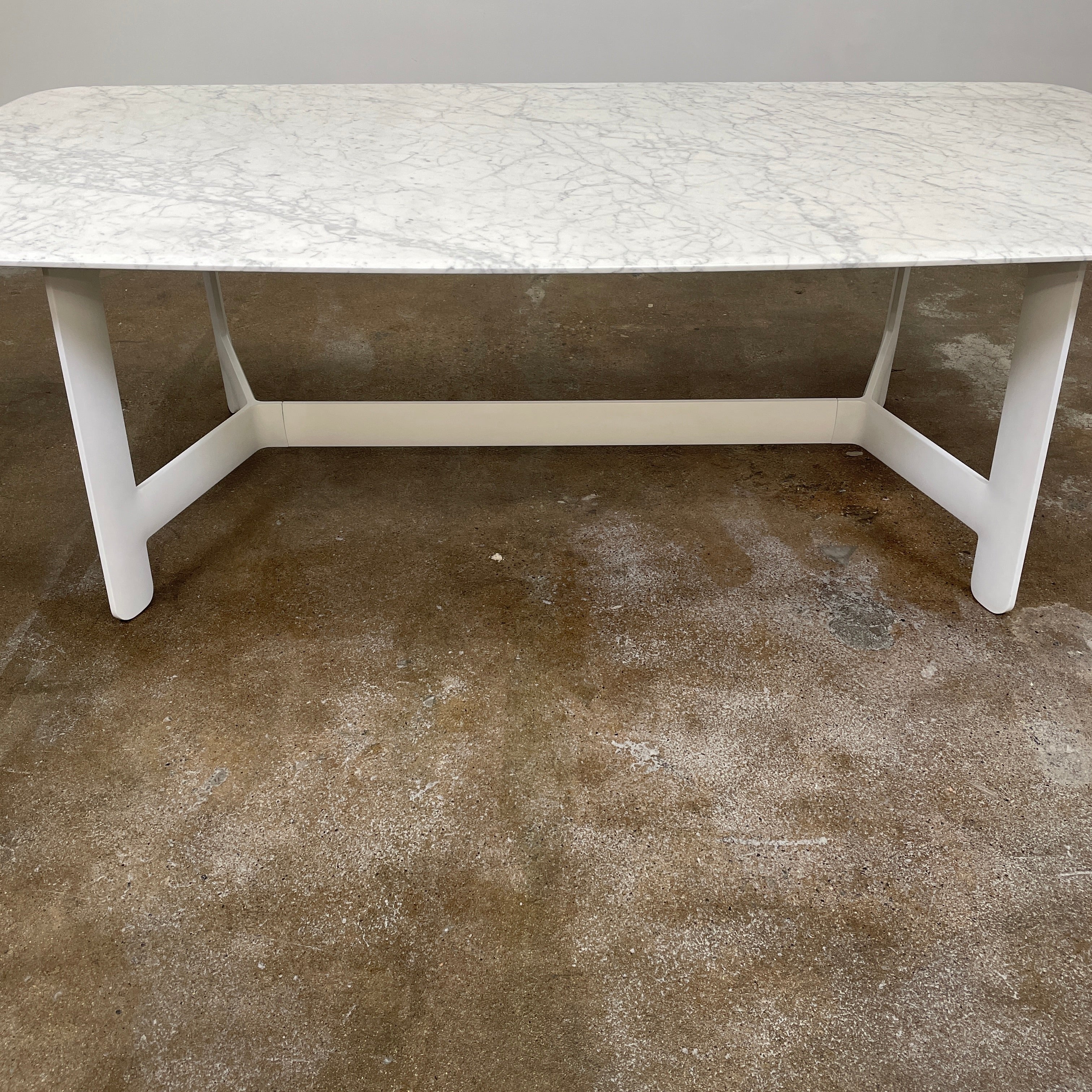 On a concrete floor against a plain white wall stands the B&B Italia Matte Carrara Marble Alex Dining Table, showcasing contemporary design with its long, rectangular form and white base, adding both style and sophistication to the space.
