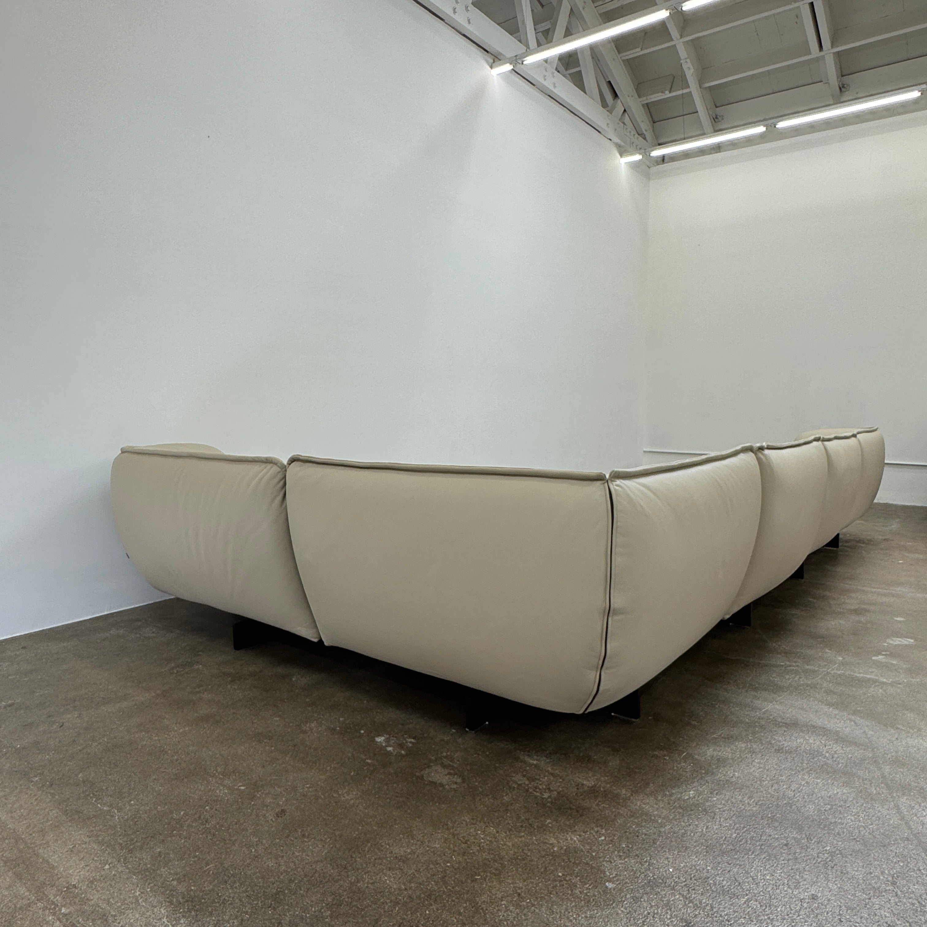 The Cassina Superbeam 5 Piece Leather Sectional in beige, featuring plush cushions, is arranged in a spacious room with white walls, exposed Superbeam ceiling, and overhead lighting.