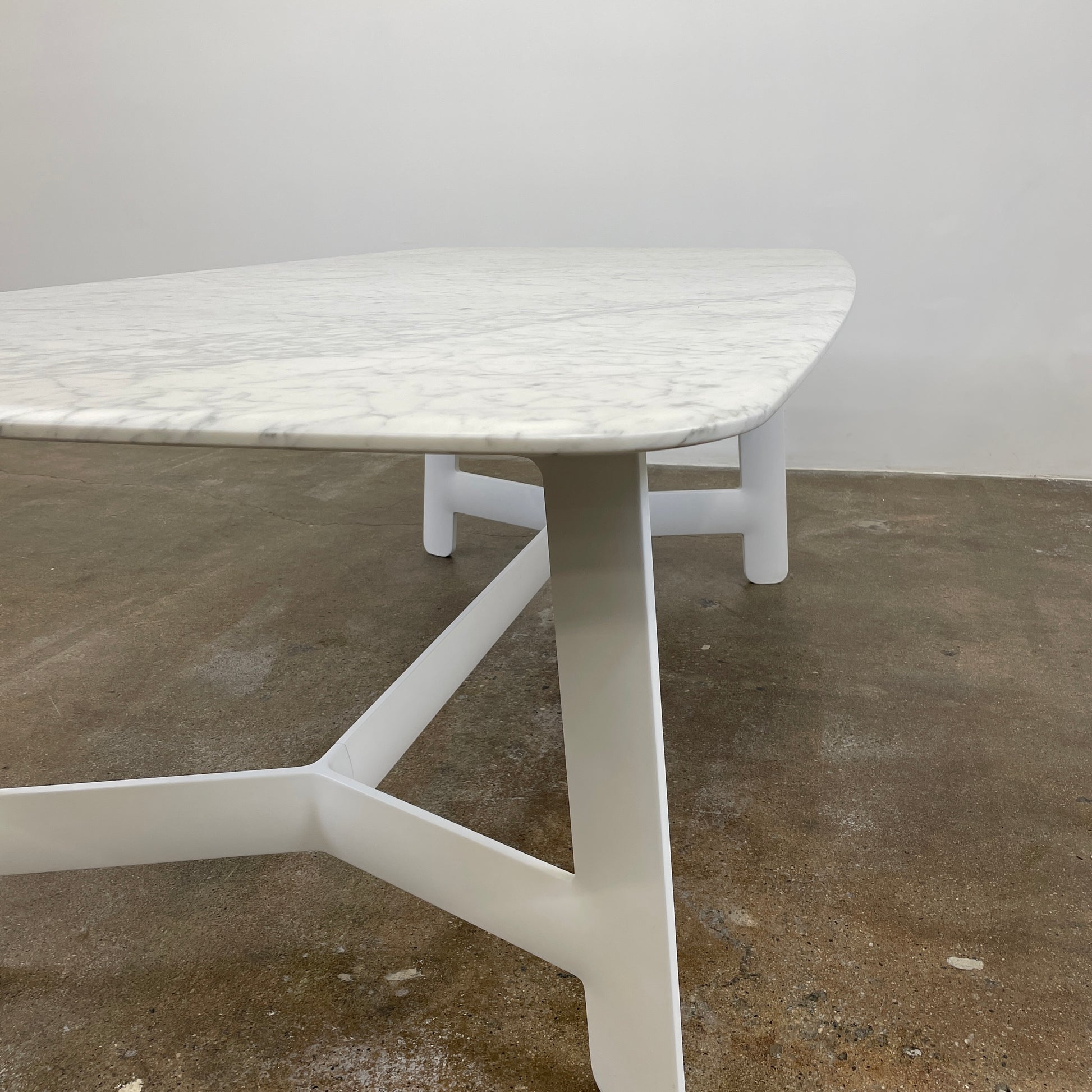 On a concrete floor against a plain white wall stands the B&B Italia Matte Carrara Marble Alex Dining Table, showcasing contemporary design with its long, rectangular form and white base, adding both style and sophistication to the space.