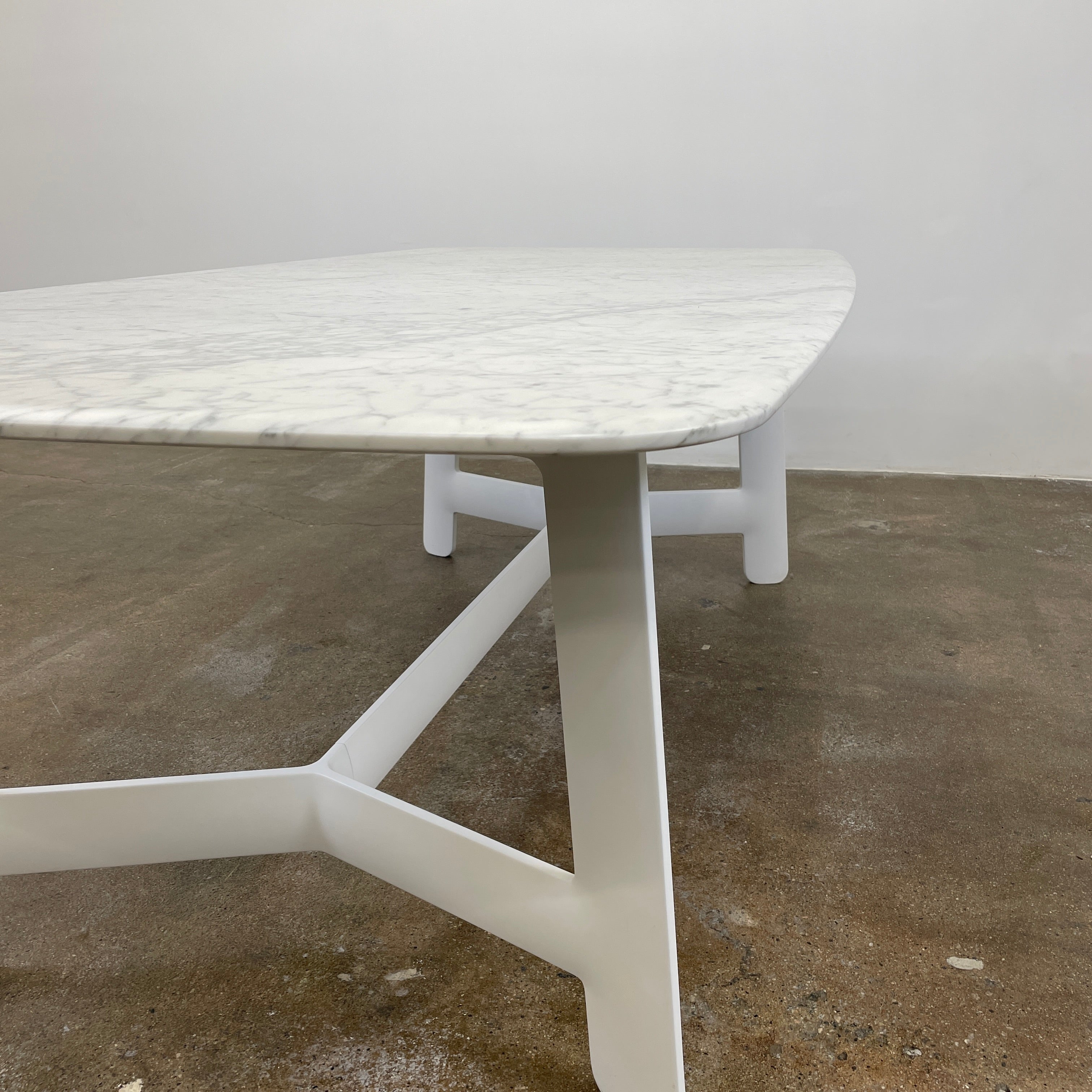 On a concrete floor against a plain white wall stands the B&B Italia Matte Carrara Marble Alex Dining Table, showcasing contemporary design with its long, rectangular form and white base, adding both style and sophistication to the space.