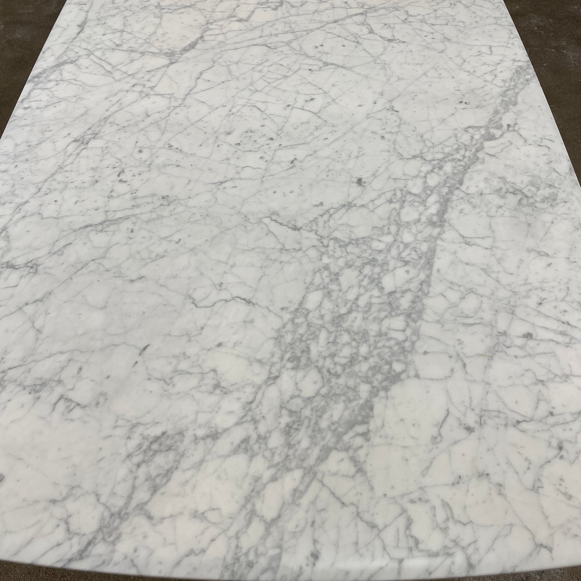 On a concrete floor against a plain white wall stands the B&B Italia Matte Carrara Marble Alex Dining Table, showcasing contemporary design with its long, rectangular form and white base, adding both style and sophistication to the space.