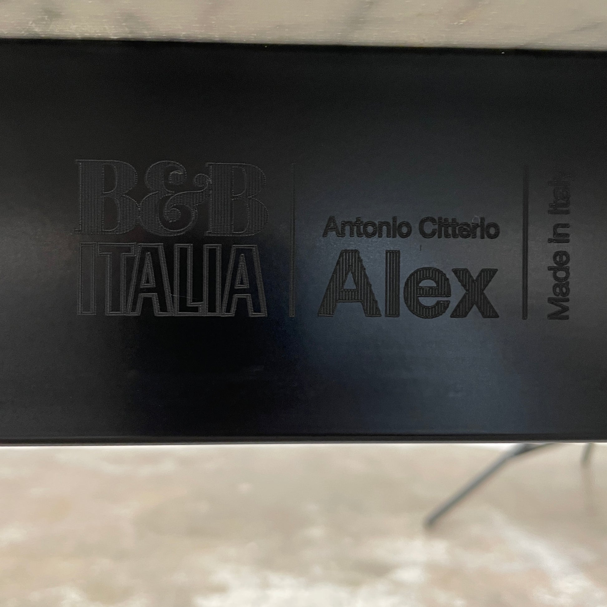 On a concrete floor against a plain white wall stands the B&B Italia Matte Carrara Marble Alex Dining Table, showcasing contemporary design with its long, rectangular form and white base, adding both style and sophistication to the space.