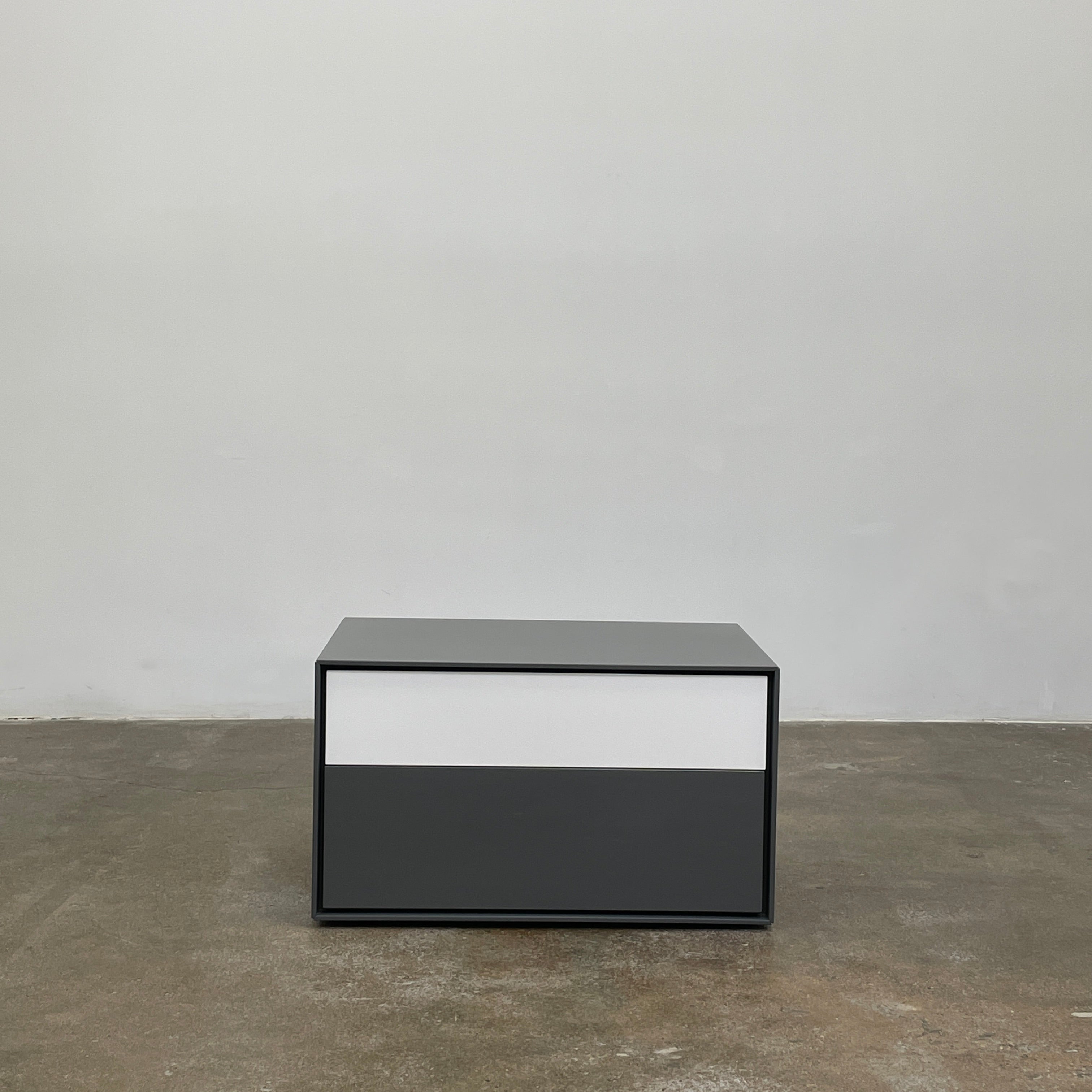 A rectangular gray and white lacquer box from B&B Italia, reminiscent of the Dado Night Table's minimalist design, features a glossy upper and matte lower section. This sleek piece serves as a stylish bedside companion, enhancing any modern space against a plain white background.