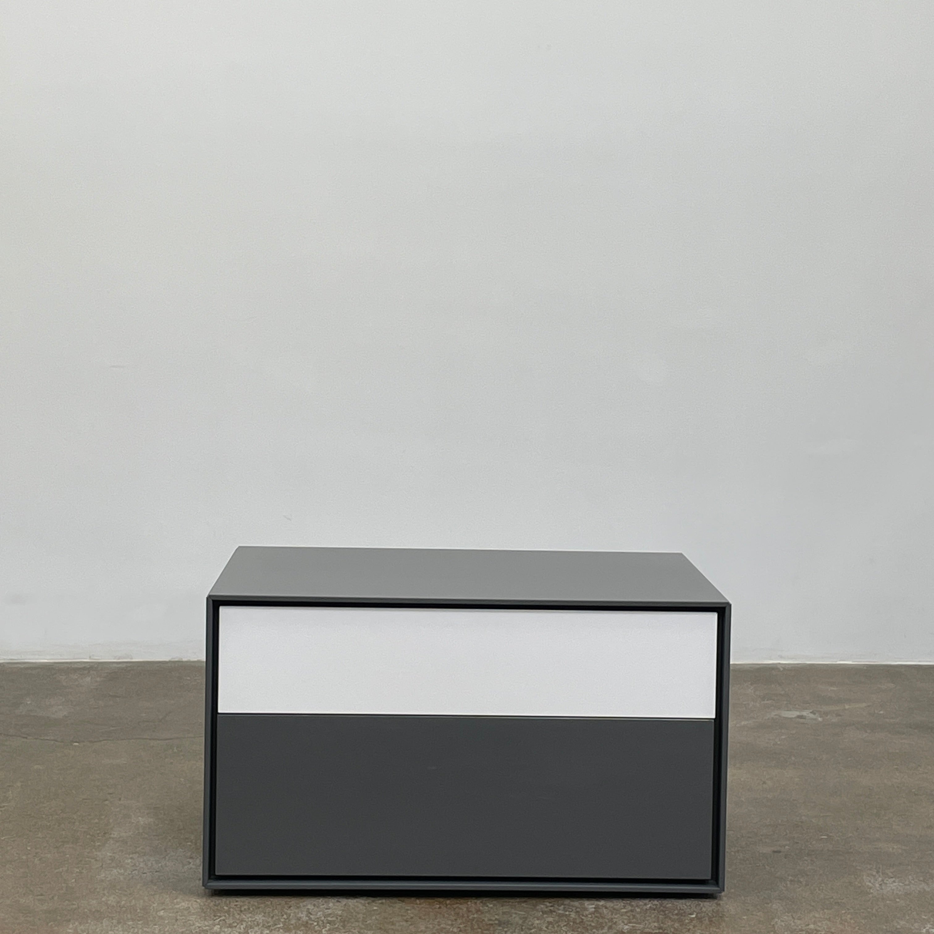 A rectangular gray and white lacquer box from B&B Italia, reminiscent of the Dado Night Table's minimalist design, features a glossy upper and matte lower section. This sleek piece serves as a stylish bedside companion, enhancing any modern space against a plain white background.