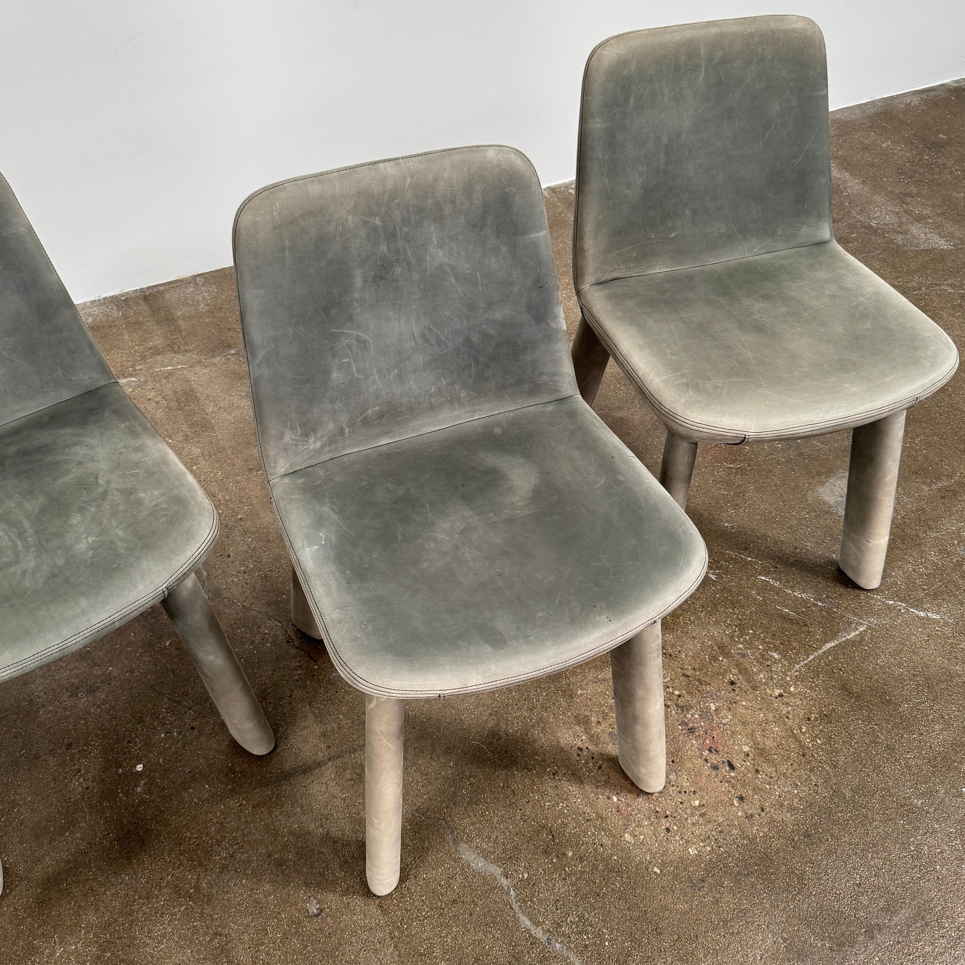 Four Blu Dot Neat Dining Chairs (set of 4, gray) from Blu Dot are lined up against a white wall on a brown concrete floor, highlighting a minimalist and tidy atmosphere.