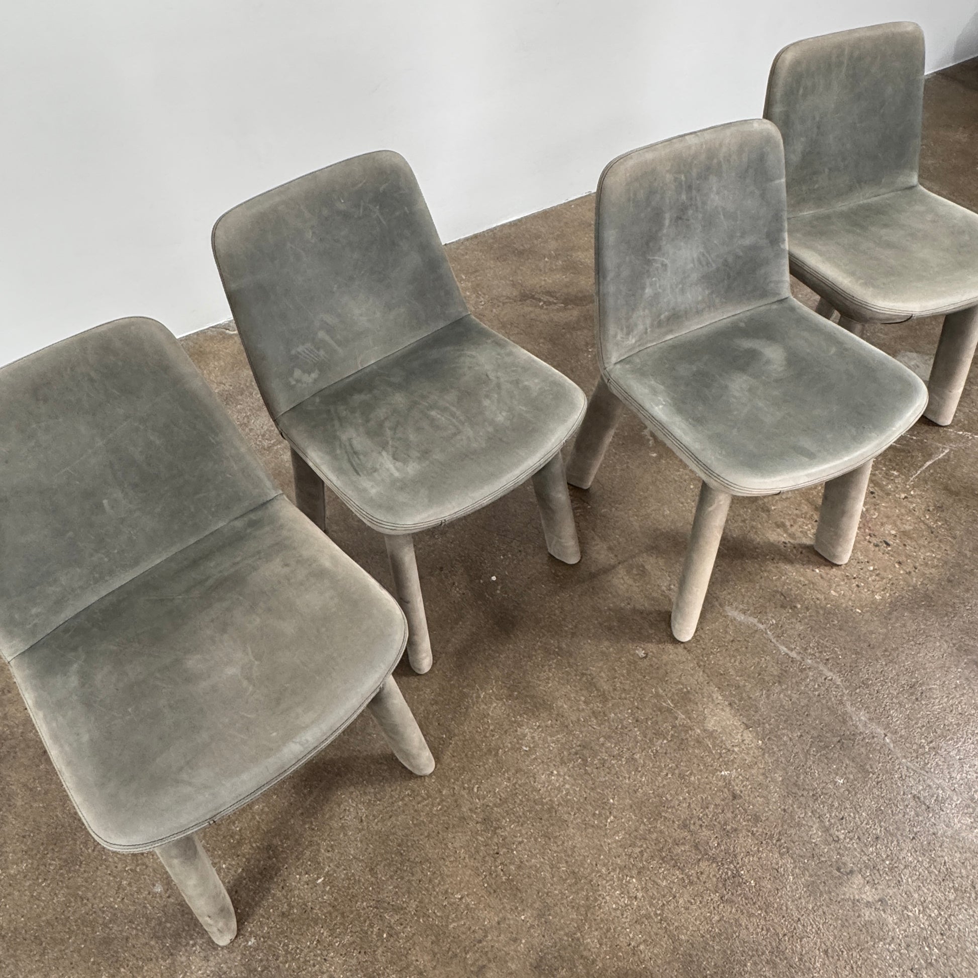 Four Blu Dot Neat Dining Chairs (set of 4, gray) from Blu Dot are lined up against a white wall on a brown concrete floor, highlighting a minimalist and tidy atmosphere.