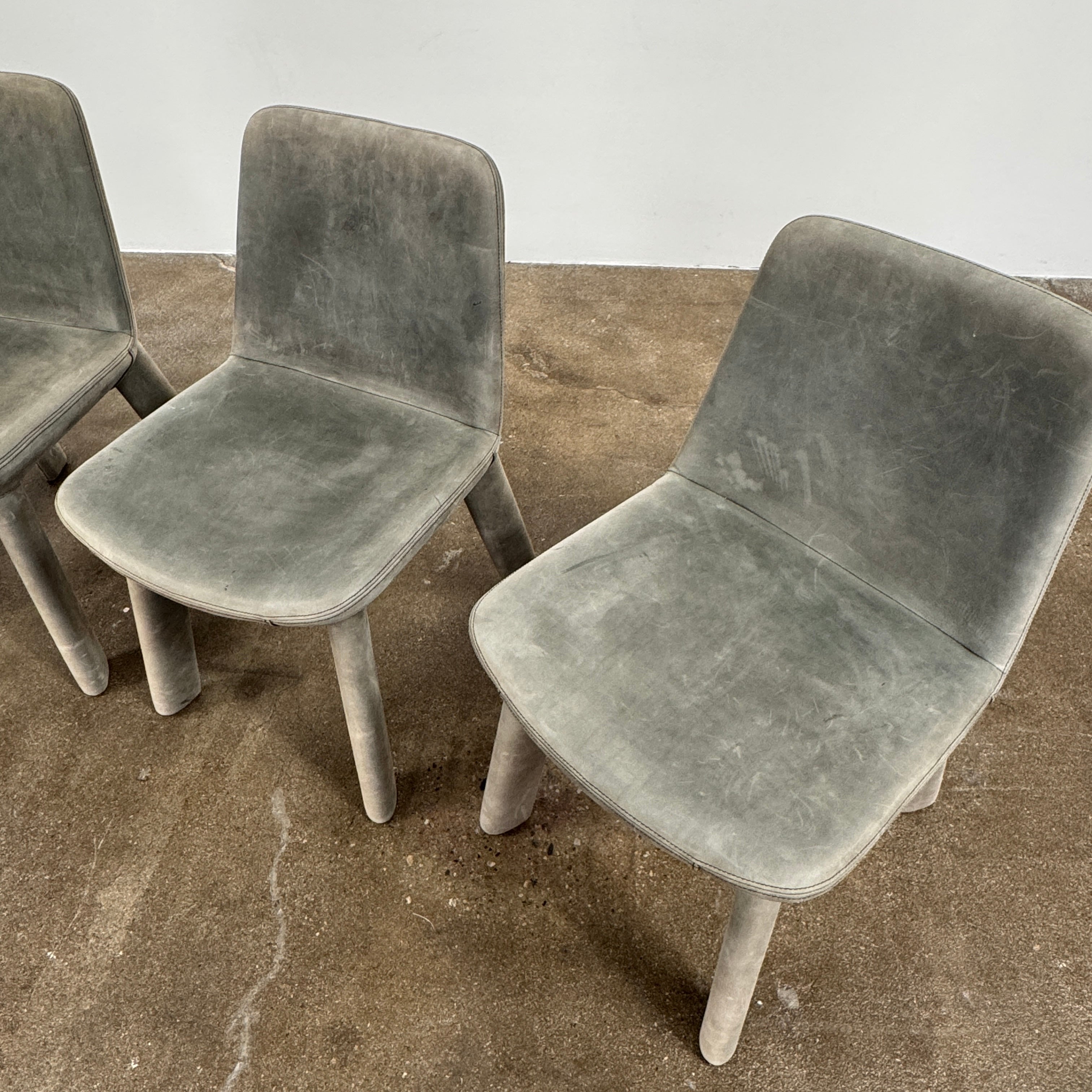 Four Blu Dot Neat Dining Chairs (set of 4, gray) from Blu Dot are lined up against a white wall on a brown concrete floor, highlighting a minimalist and tidy atmosphere.