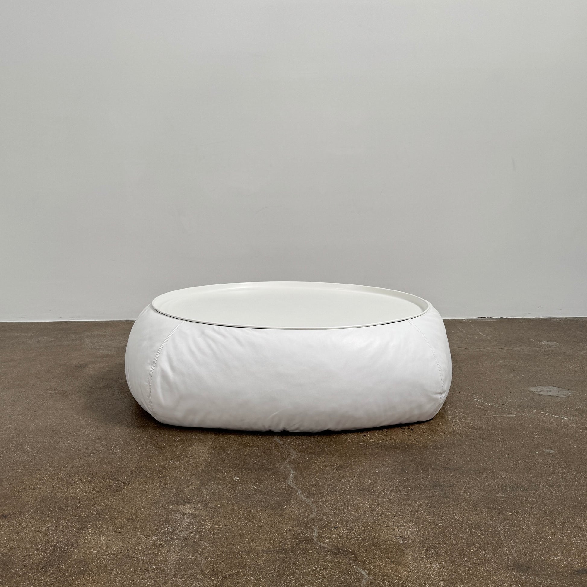 The B&B Italia Lady Fat Coffee Table by B&B Italia, a low, round table with a flat white top inspired by Patricia Urquiola’s design, sits on a plain brown floor against a light gray wall.