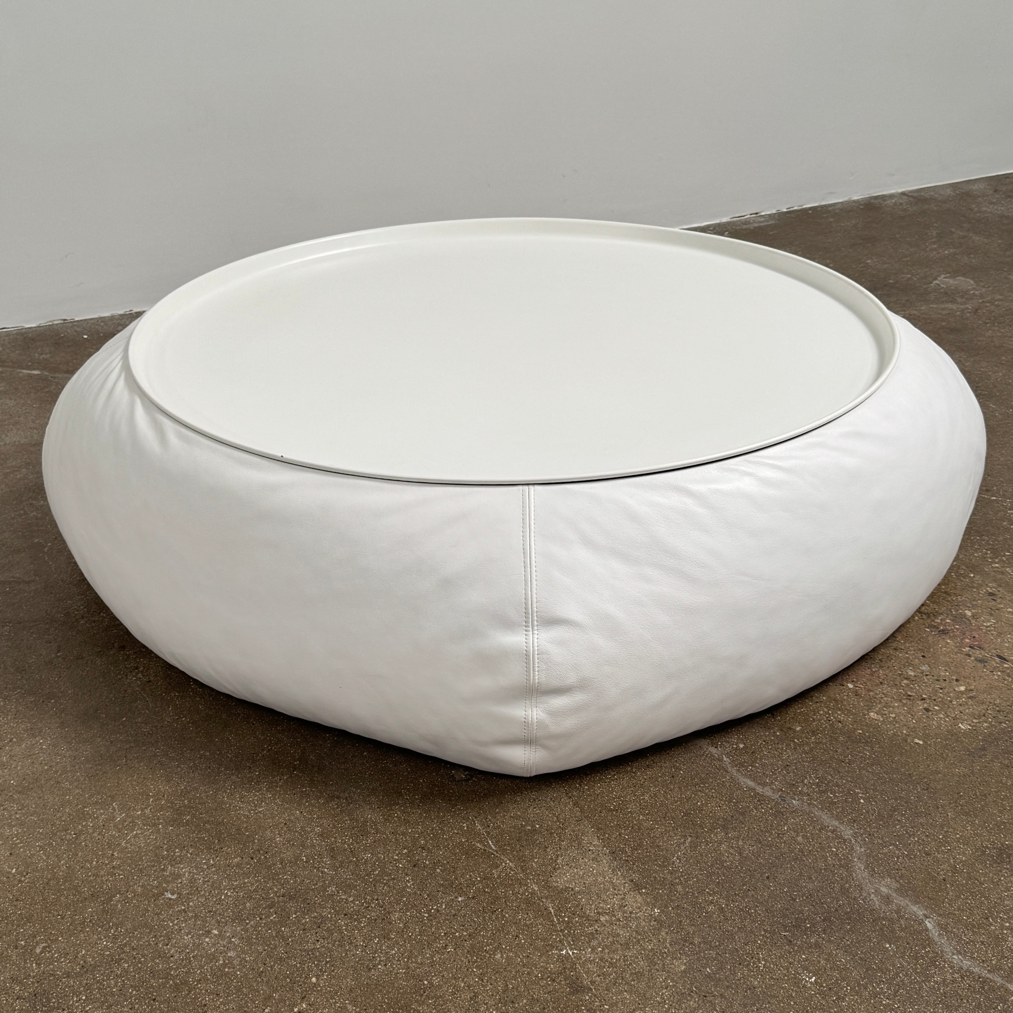 The B&B Italia Lady Fat Coffee Table by B&B Italia, a low, round table with a flat white top inspired by Patricia Urquiola’s design, sits on a plain brown floor against a light gray wall.
