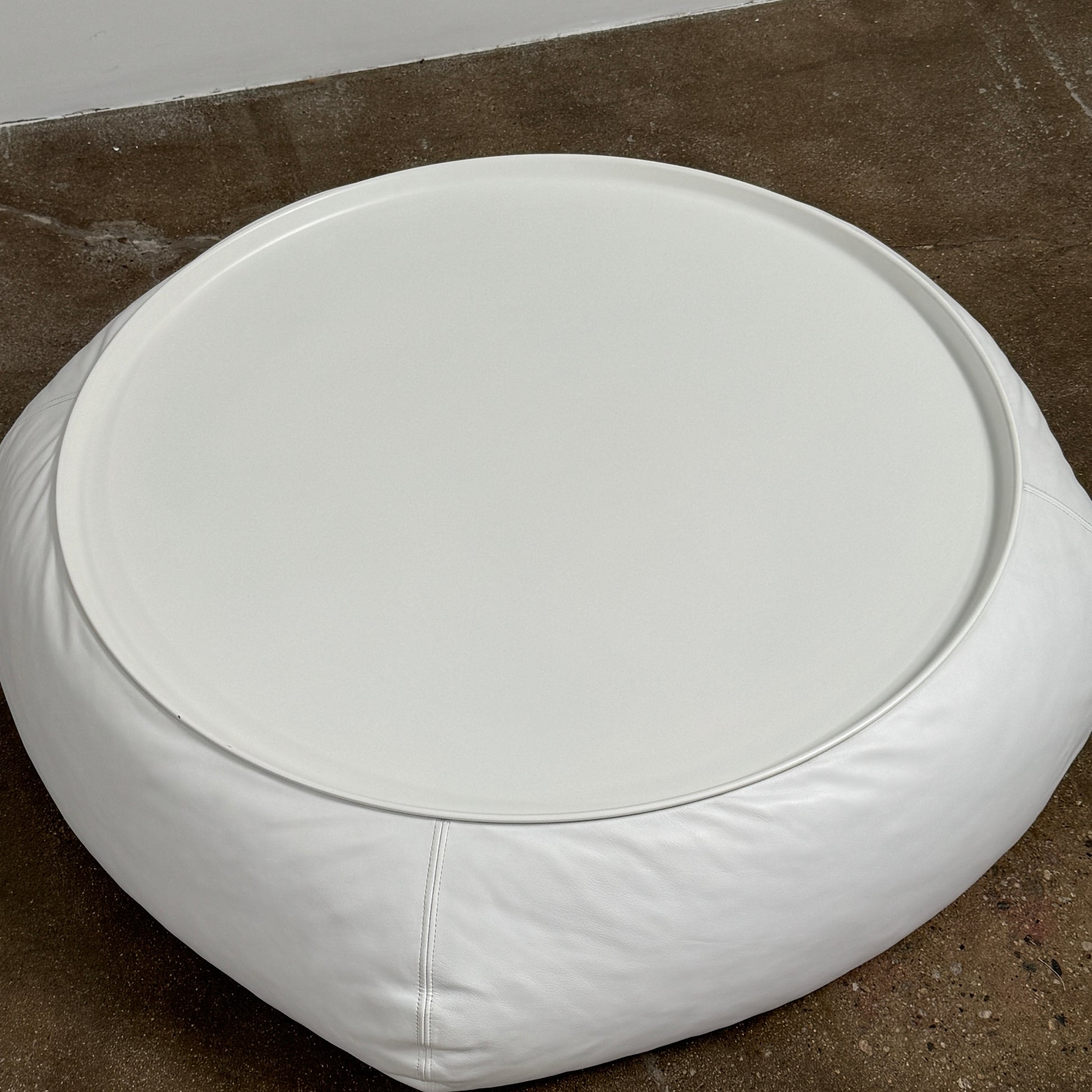 The B&B Italia Lady Fat Coffee Table by B&B Italia, a low, round table with a flat white top inspired by Patricia Urquiola’s design, sits on a plain brown floor against a light gray wall.