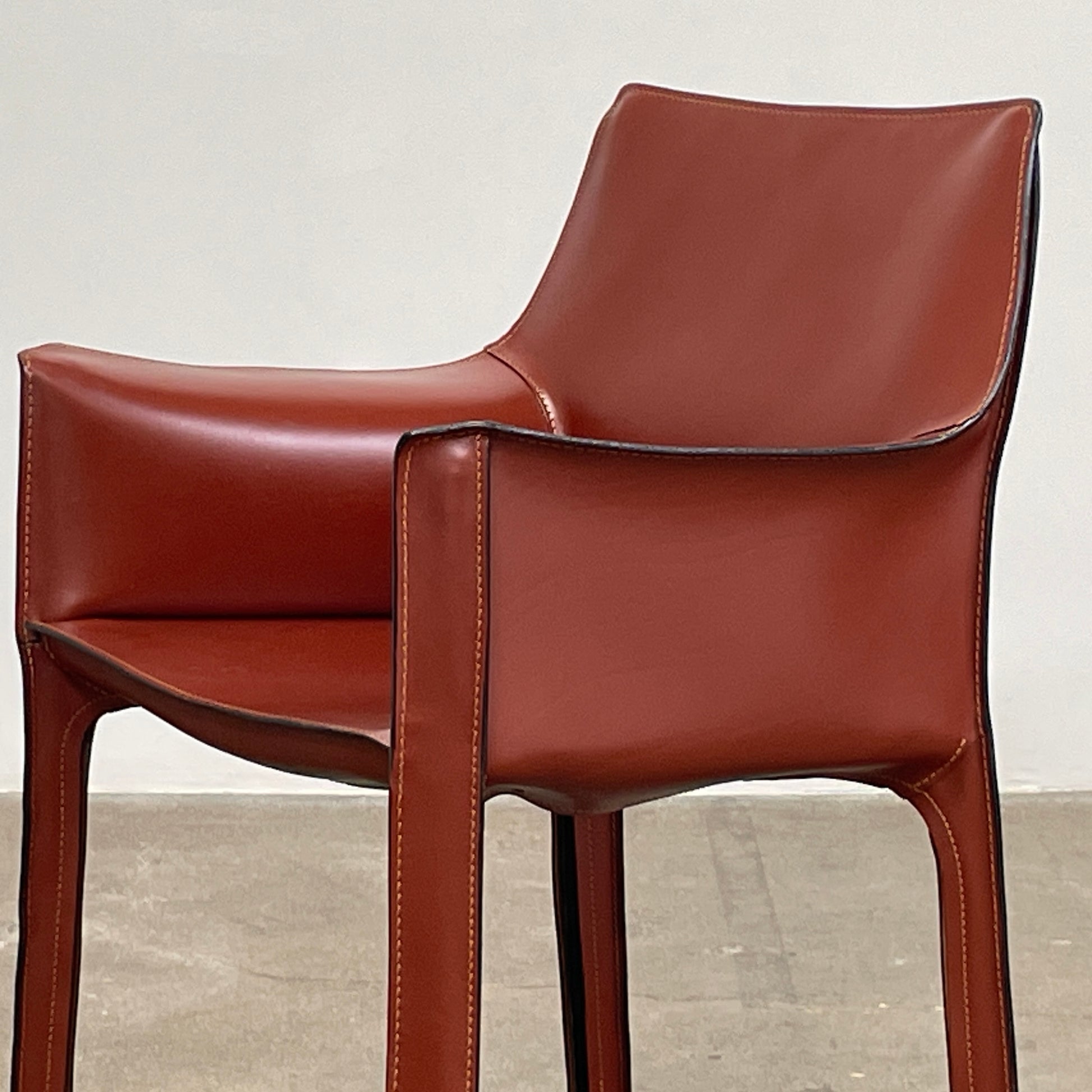 A red leather armchair, the Cassina Cab 413, drawing inspiration from the iconic Cab Chair with clean lines and a minimalist design, set against a plain grey background.