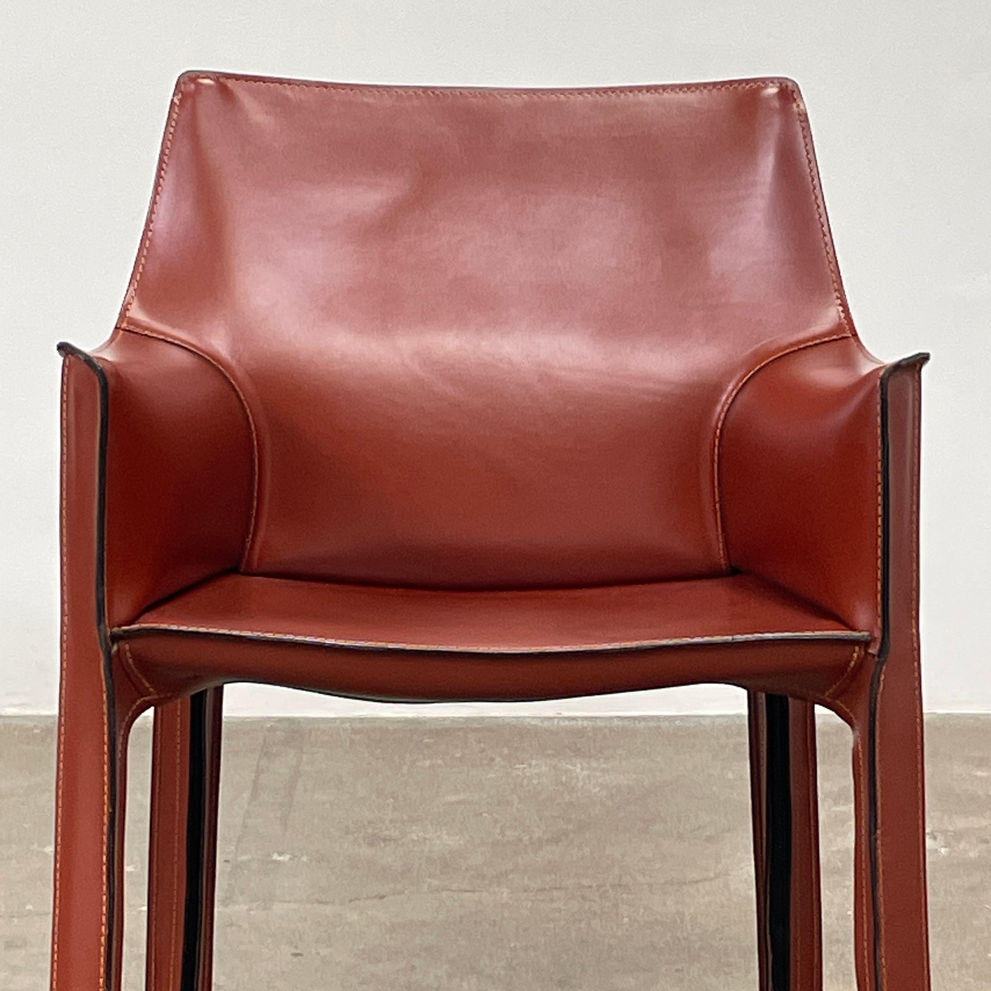 A red leather armchair, the Cassina Cab 413, drawing inspiration from the iconic Cab Chair with clean lines and a minimalist design, set against a plain grey background.