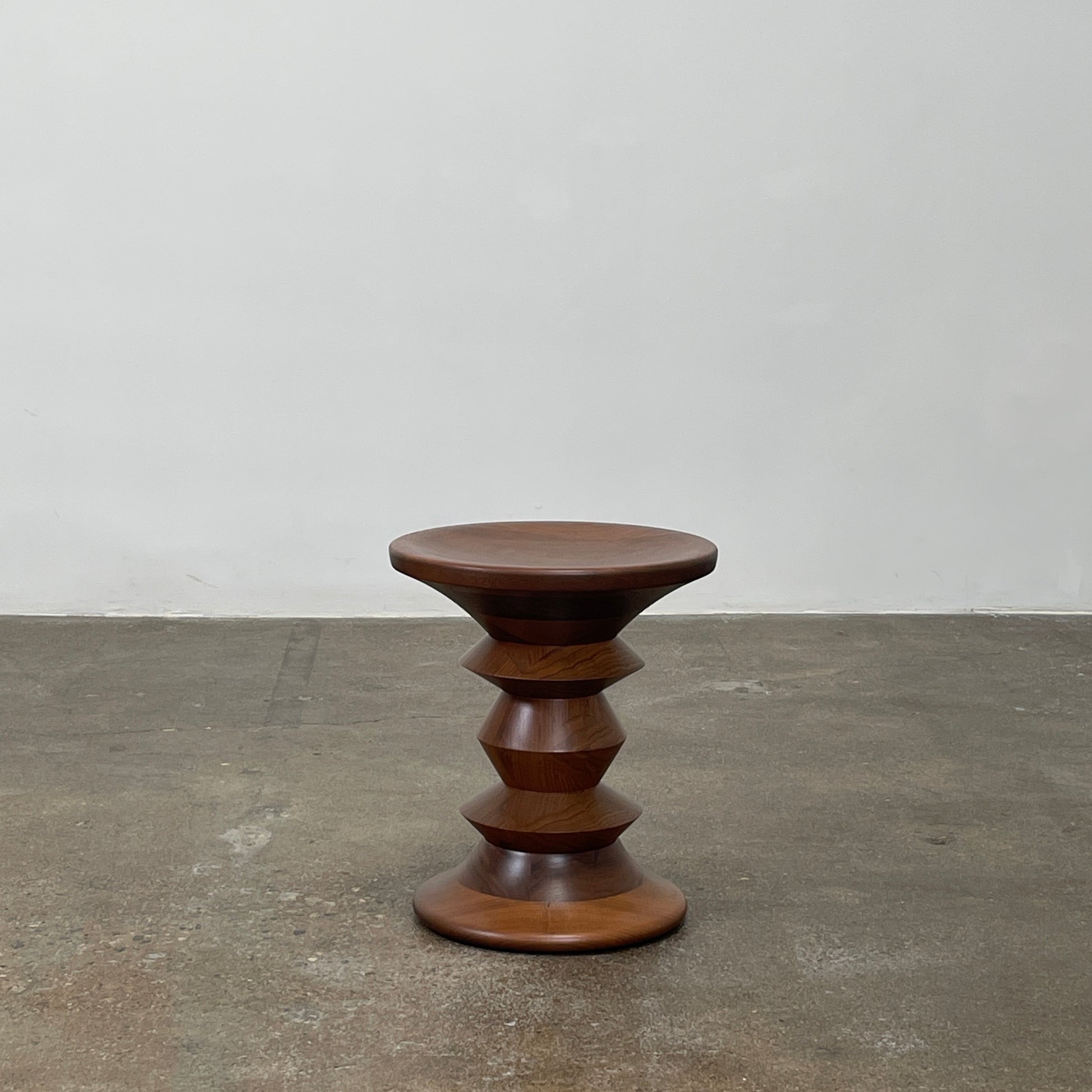 The Eames Walnut Turned Shape C Stool by Herman Miller, featuring a solid-turned walnut design with a base resembling an abstract chess piece, stands on a concrete floor against a plain white wall.