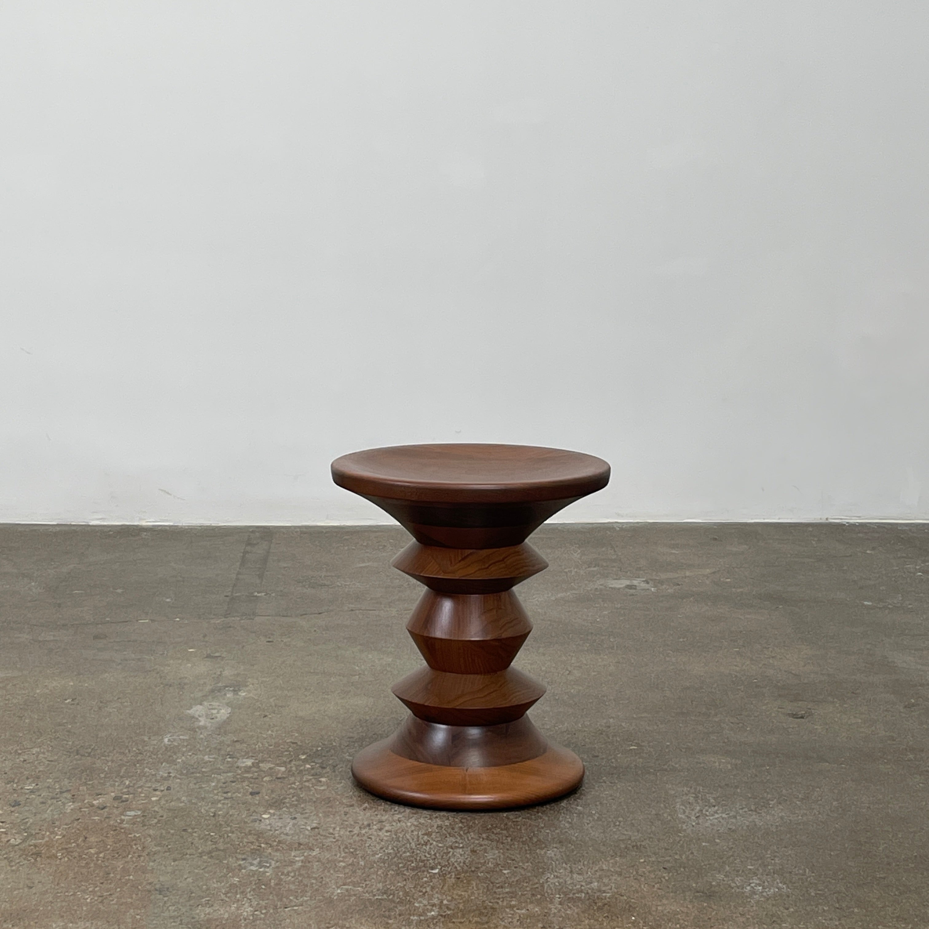 The Eames Walnut Turned Shape C Stool by Herman Miller, featuring a solid-turned walnut design with a base resembling an abstract chess piece, stands on a concrete floor against a plain white wall.