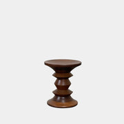 The Eames Walnut Turned Shape C Stool by Herman Miller, featuring a solid-turned walnut design with a base resembling an abstract chess piece, stands on a concrete floor against a plain white wall.