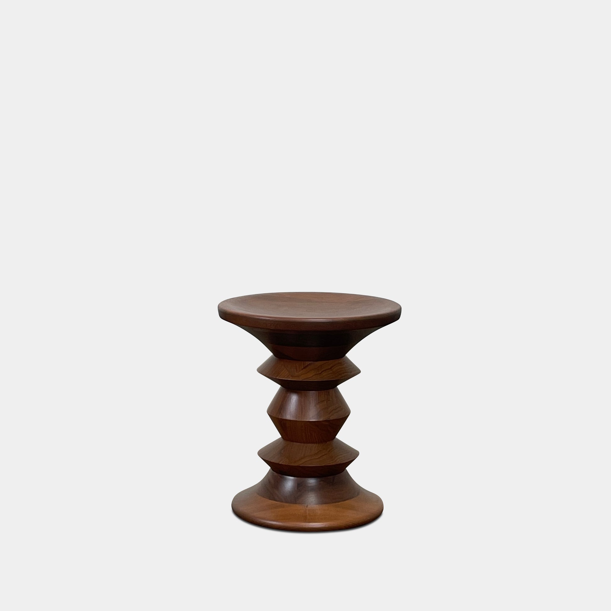 The Eames Walnut Turned Shape C Stool by Herman Miller, featuring a solid-turned walnut design with a base resembling an abstract chess piece, stands on a concrete floor against a plain white wall.