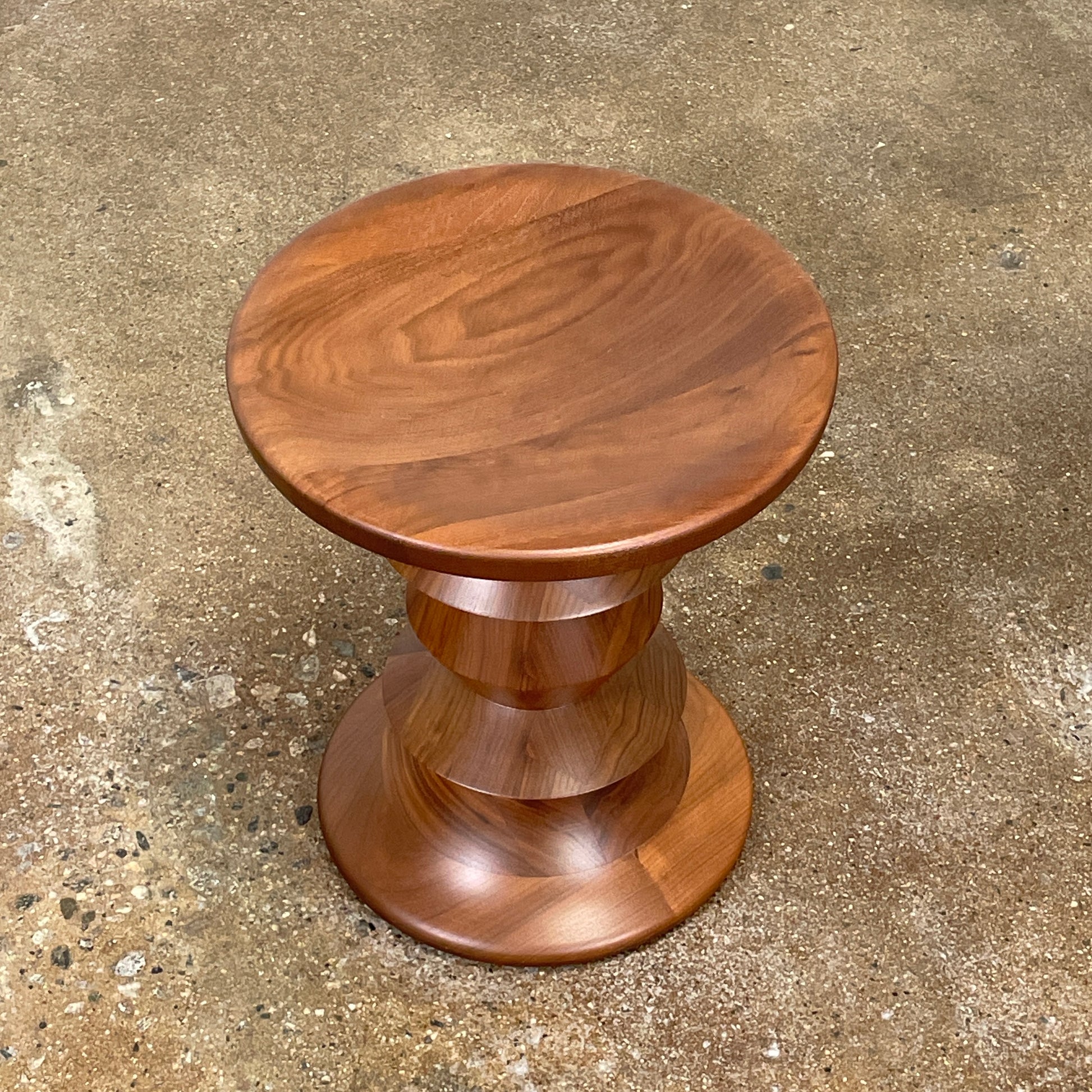 The Eames Walnut Turned Shape C Stool by Herman Miller, featuring a solid-turned walnut design with a base resembling an abstract chess piece, stands on a concrete floor against a plain white wall.