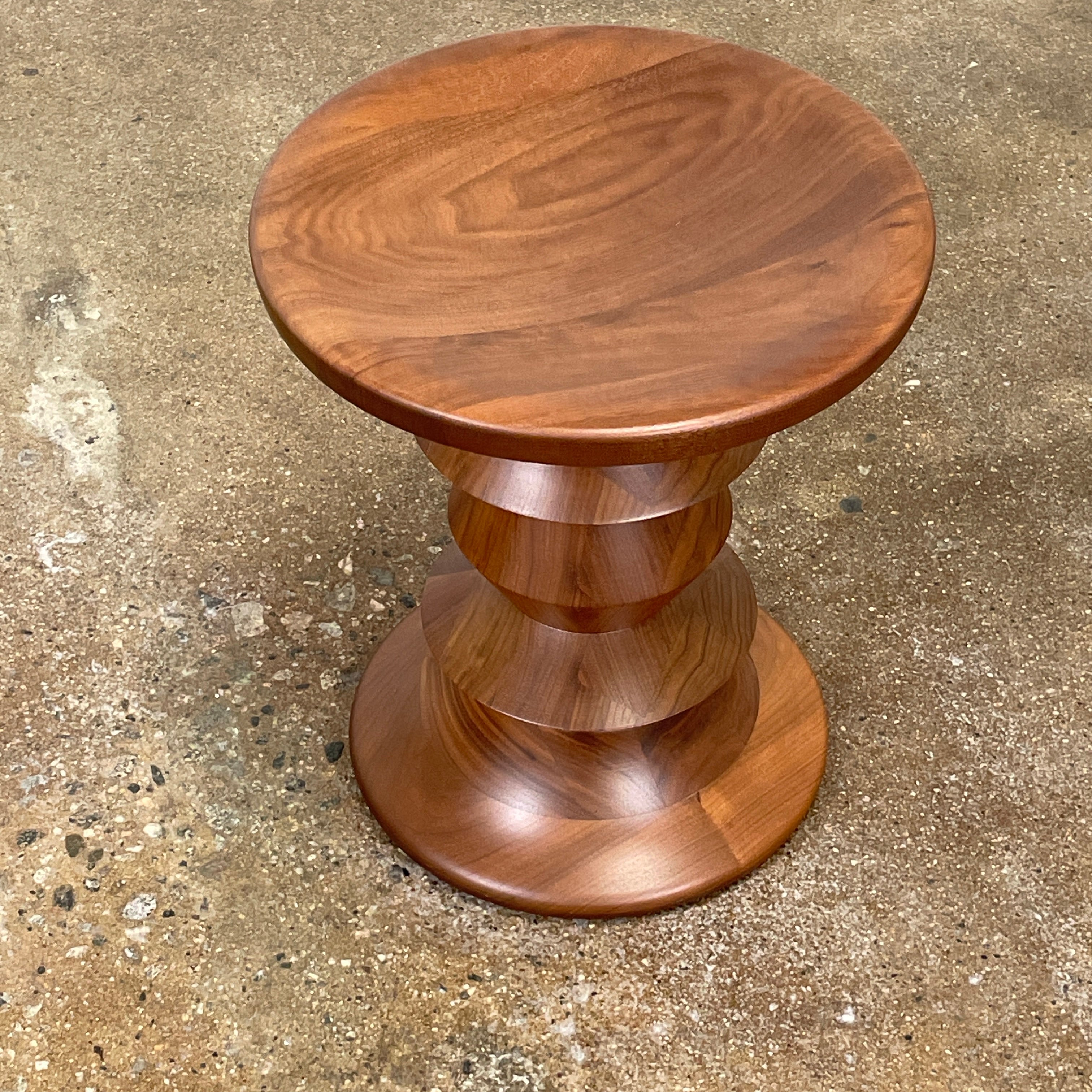 The Eames Walnut Turned Shape C Stool by Herman Miller, featuring a solid-turned walnut design with a base resembling an abstract chess piece, stands on a concrete floor against a plain white wall.