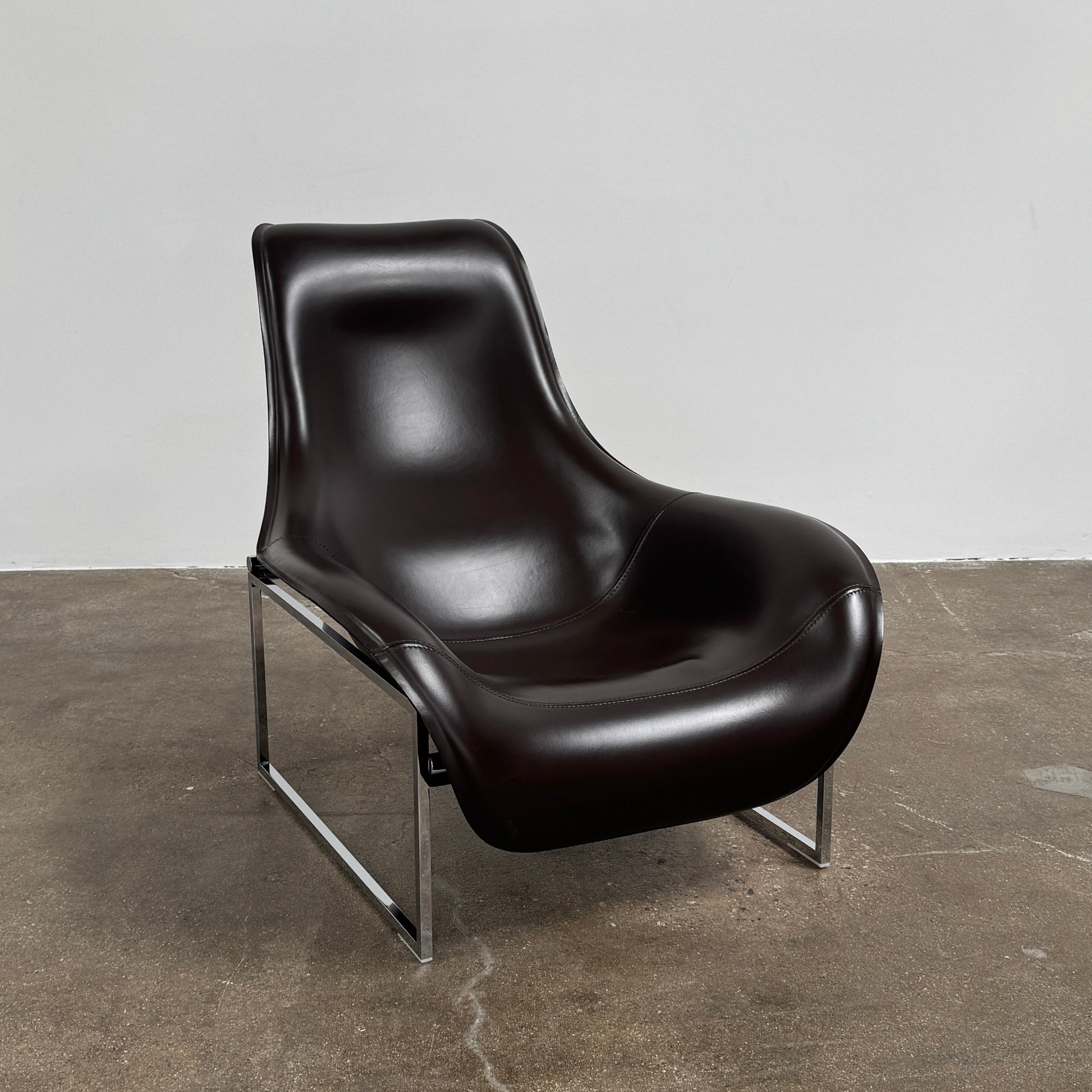 A B&B Italia Mart Relax Armchair with Tilting Mechanism in black saddle leather features a modern, curved design and is set on a polished concrete floor against a plain light gray wall.