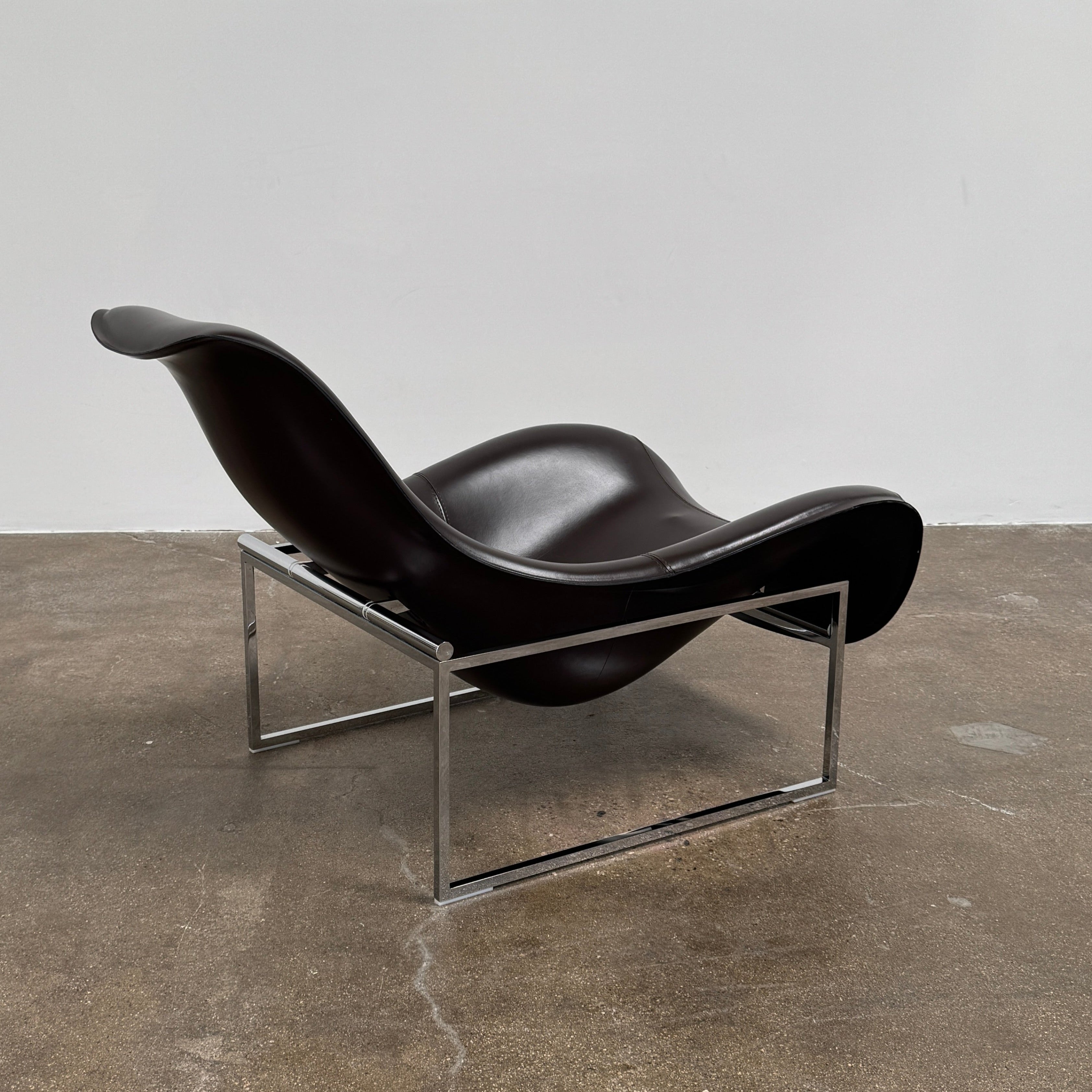 A B&B Italia Mart Relax Armchair with Tilting Mechanism in black saddle leather features a modern, curved design and is set on a polished concrete floor against a plain light gray wall.