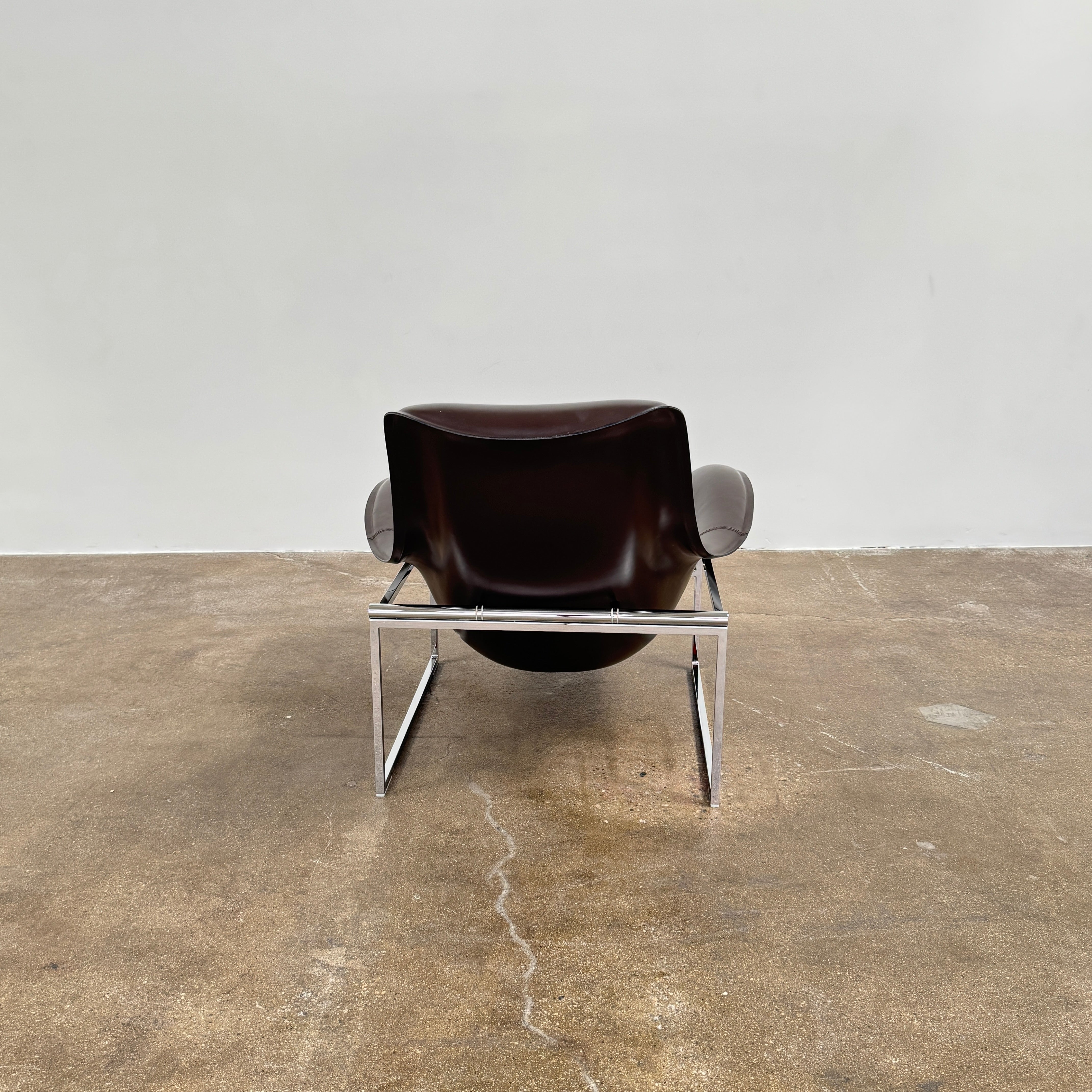 A B&B Italia Mart Relax Armchair with Tilting Mechanism in black saddle leather features a modern, curved design and is set on a polished concrete floor against a plain light gray wall.