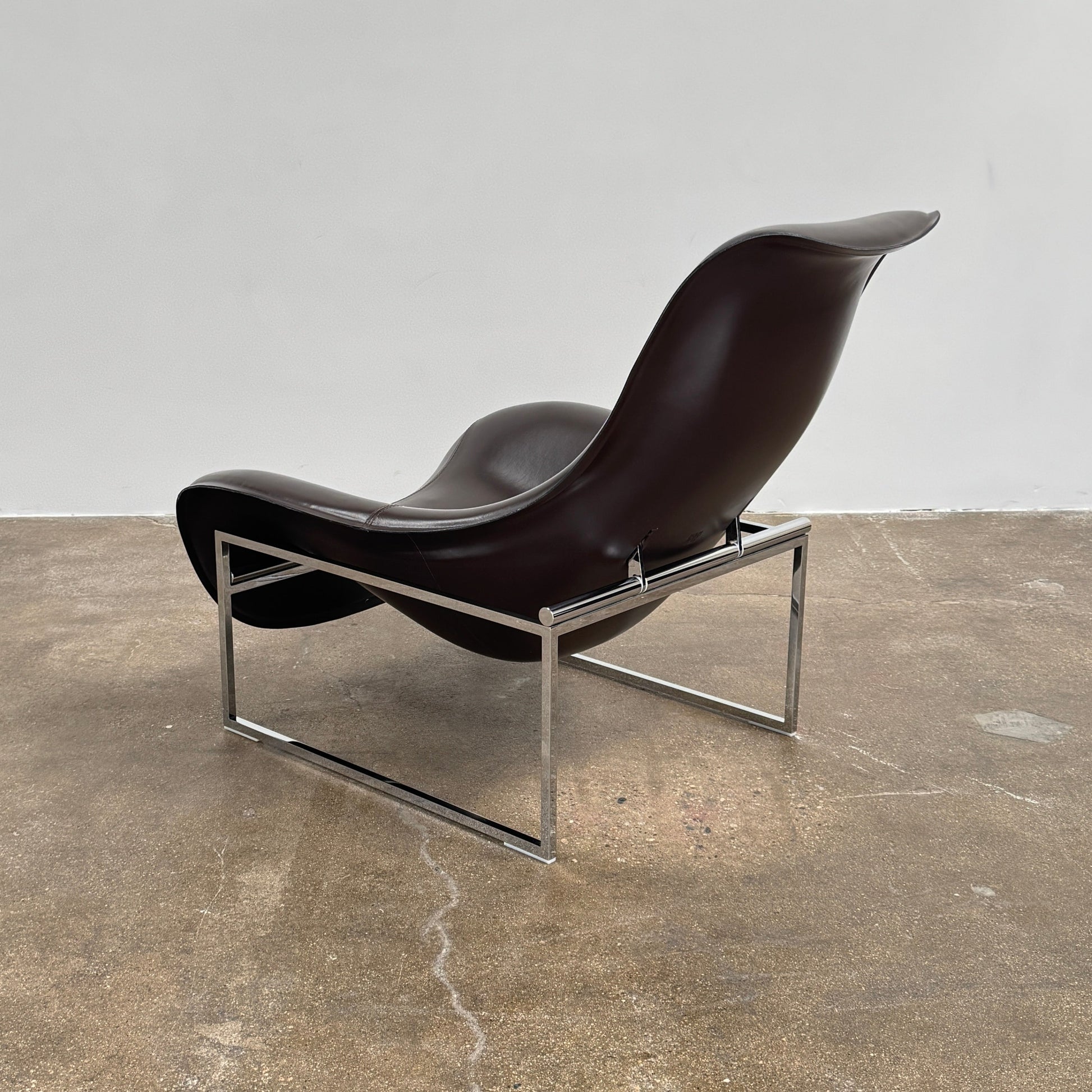 A B&B Italia Mart Relax Armchair with Tilting Mechanism in black saddle leather features a modern, curved design and is set on a polished concrete floor against a plain light gray wall.